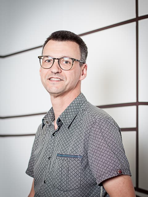 foto_eric_iminds Portret van Eric Van der Hulst met een bril en een overhemd met patroon tegen een lichte geometrische achtergrond.