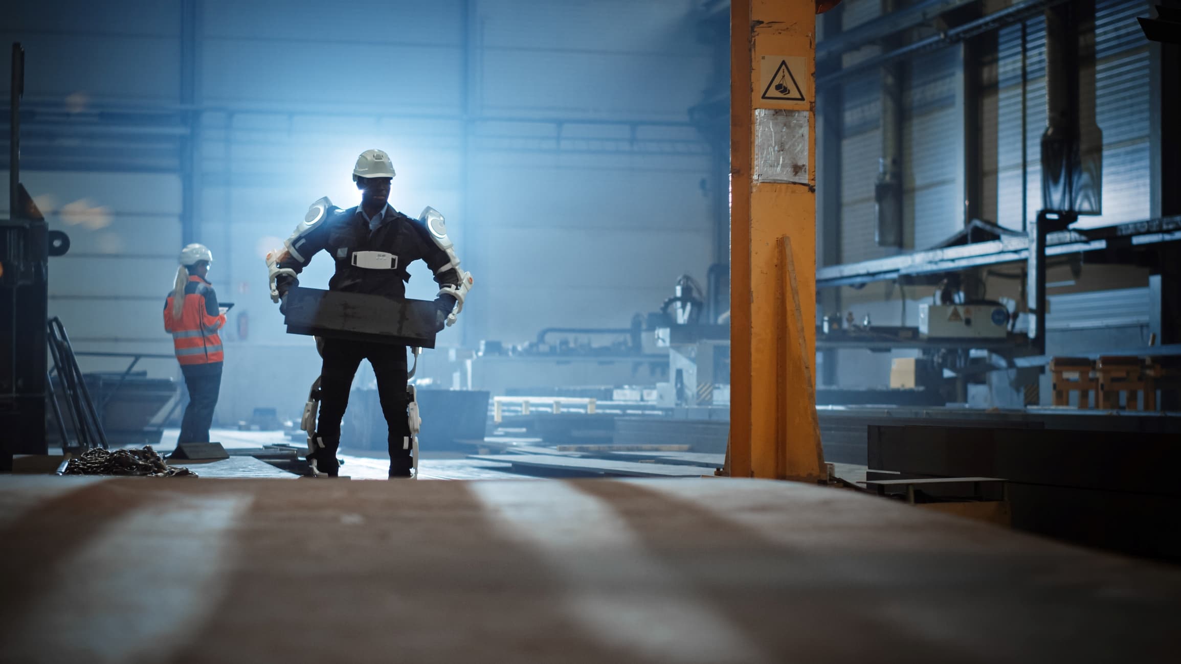 GettyImages-1290244683 Werknemer met exoskelet in een bouwhal, met een persoon in een veiligheidsvest in de buurt