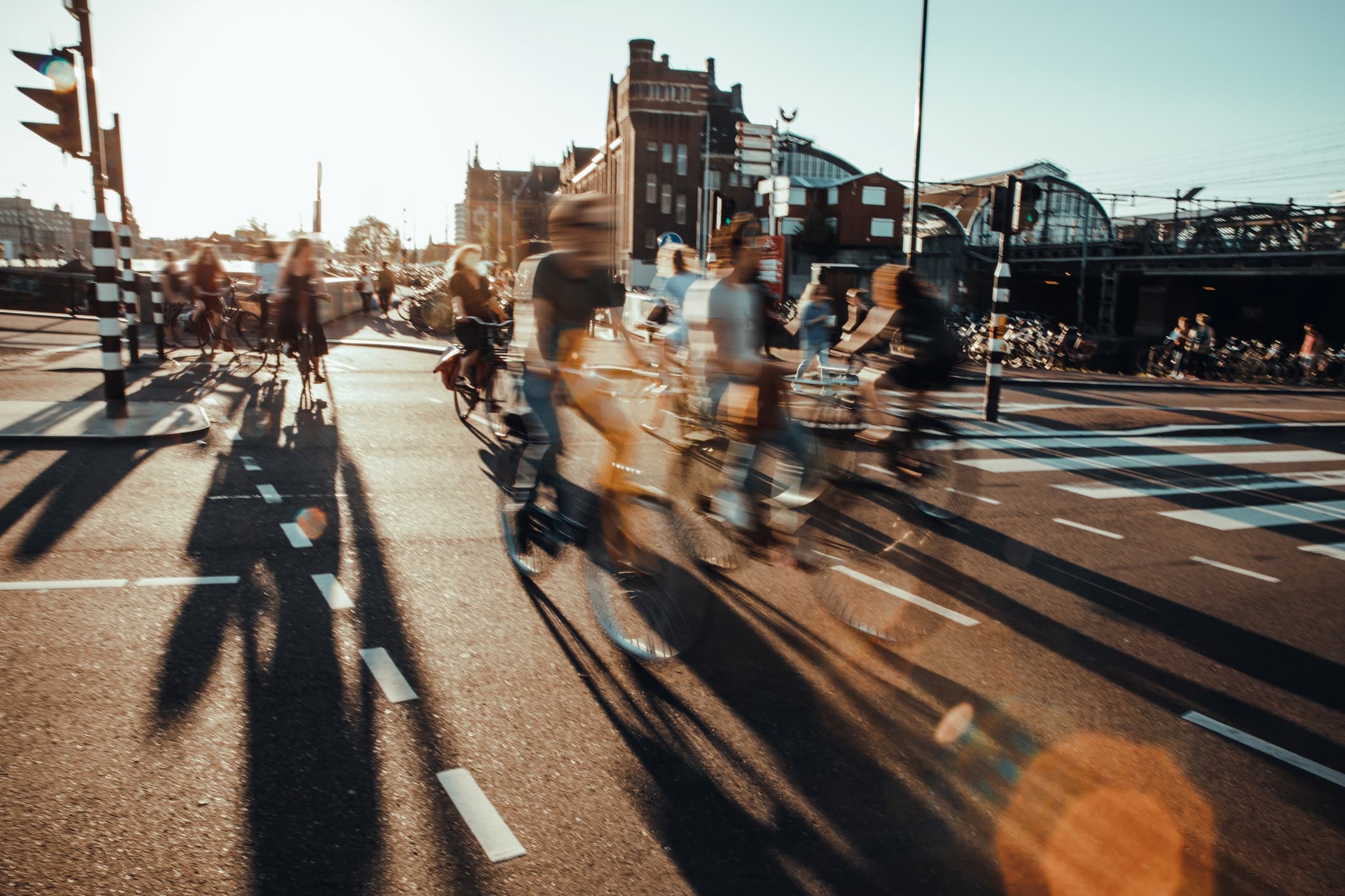 GettyImages-1361690416_0 Wazige fietsers die een druk stadsplein oversteken bij zonsondergang, met lange schaduwen en zichtbare verkeerslichten.