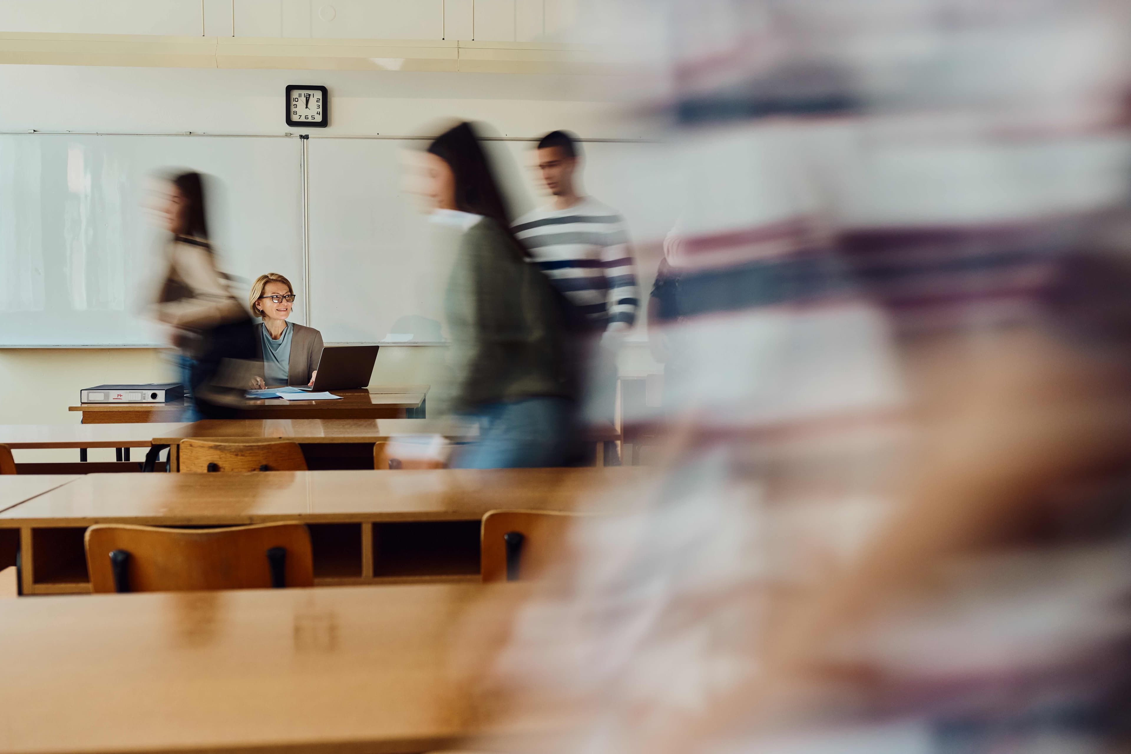 GettyImages-1819812205%20%281%29_0 Leraar aan een bureau in het klaslokaal met een laptop terwijl studenten op de voorgrond langslopen