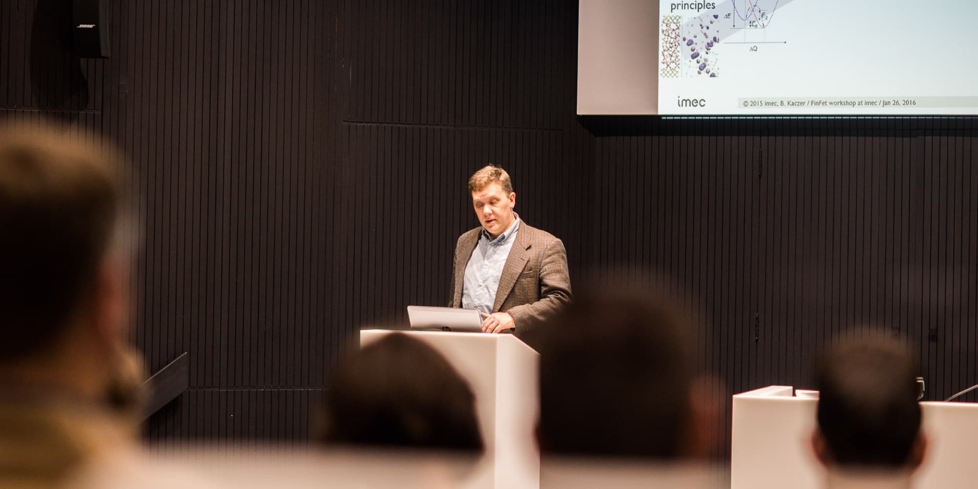 Government-funded-research Speaker presenting at a research event with a projected slide behind him and audience in the foreground