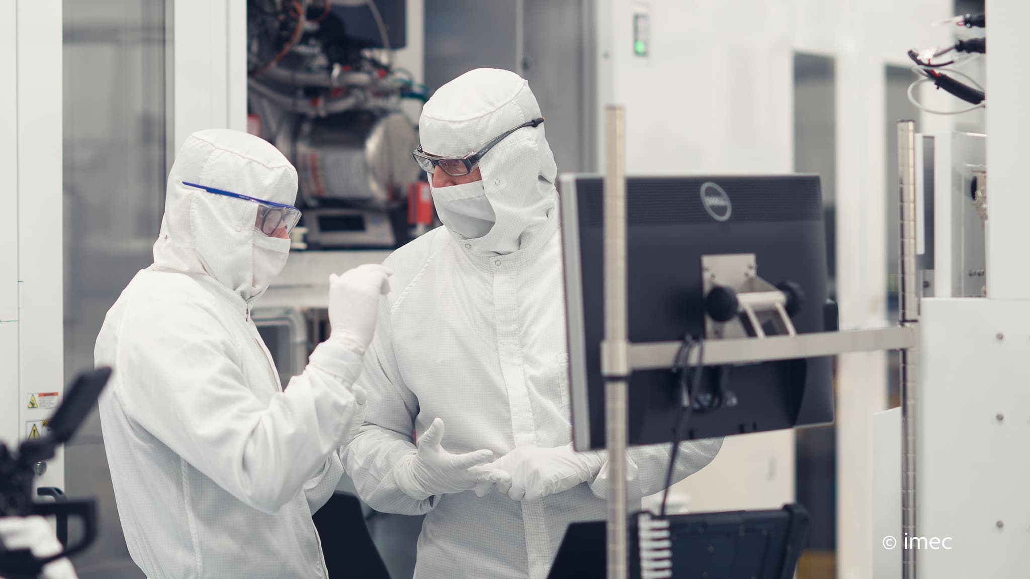 Header_0 Two cleanroom researchers in protective suits discussing work beside semiconductor equipment