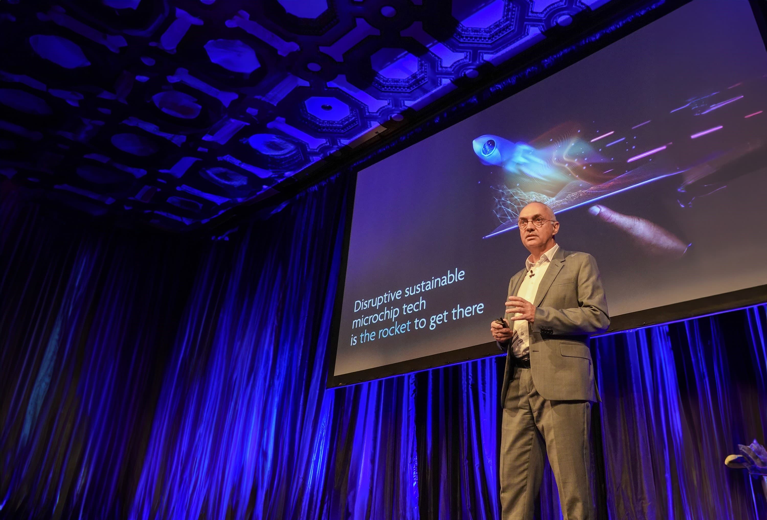 Speaker presenting sustainable microchip technology Speaker on stage beside a presentation screen about disruptive sustainable microchip tech