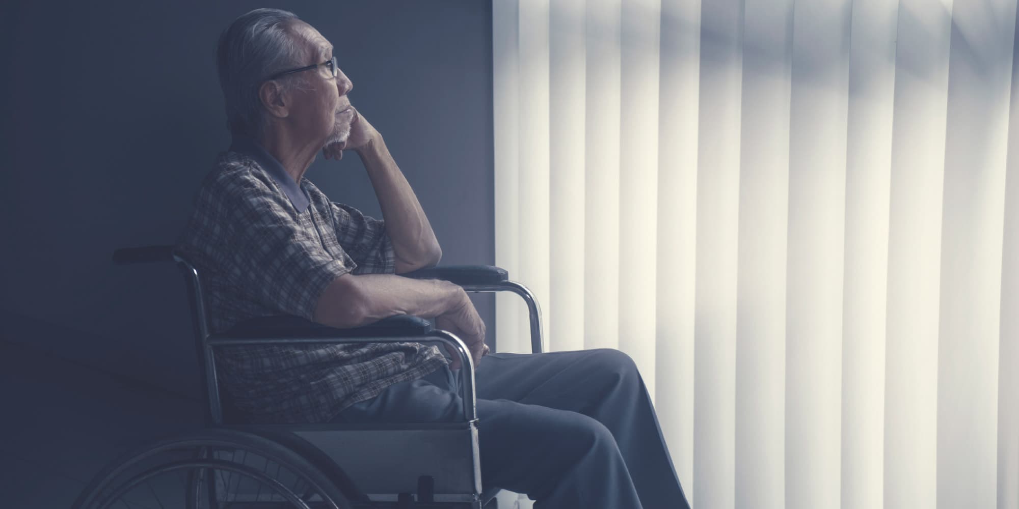 elderly man in wheelchair looking out window Oudere man in een rolstoel die naar een helder raam met verticale jaloezieën kijkt