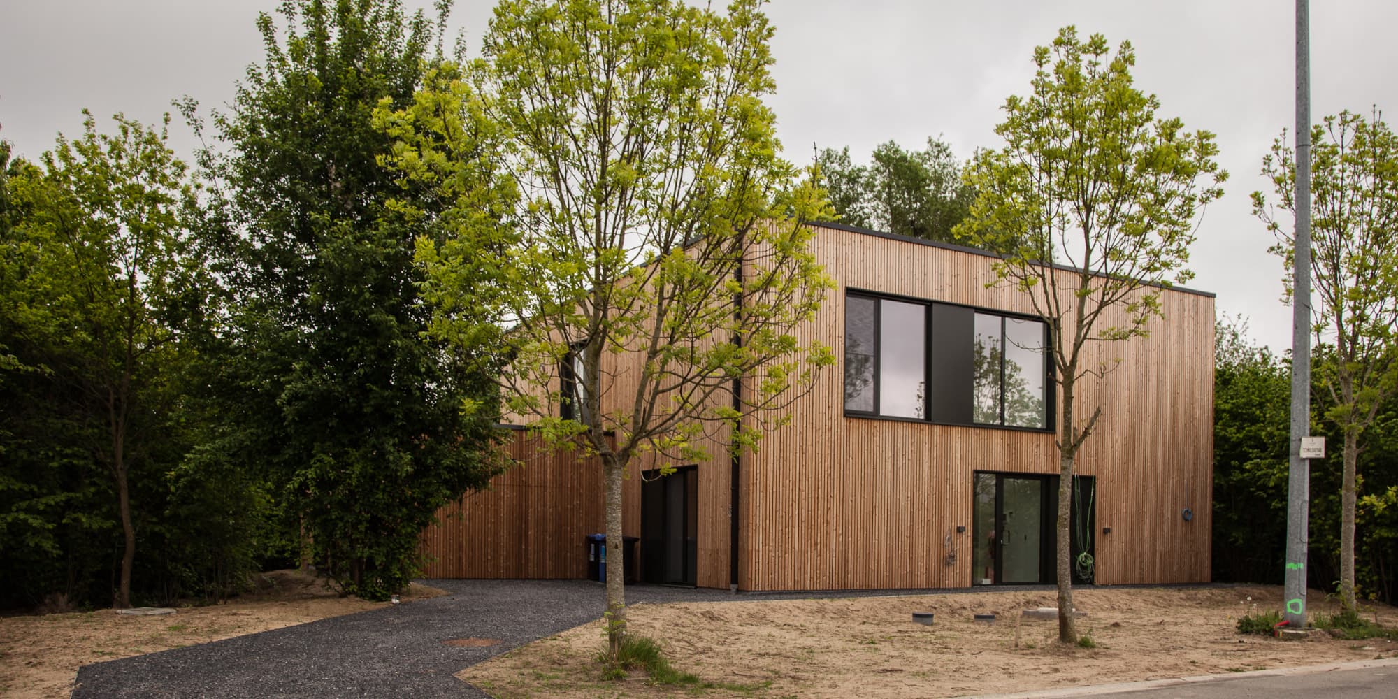 homelab_1 Modern wooden house surrounded by trees, with large black-framed windows and a gravel driveway.