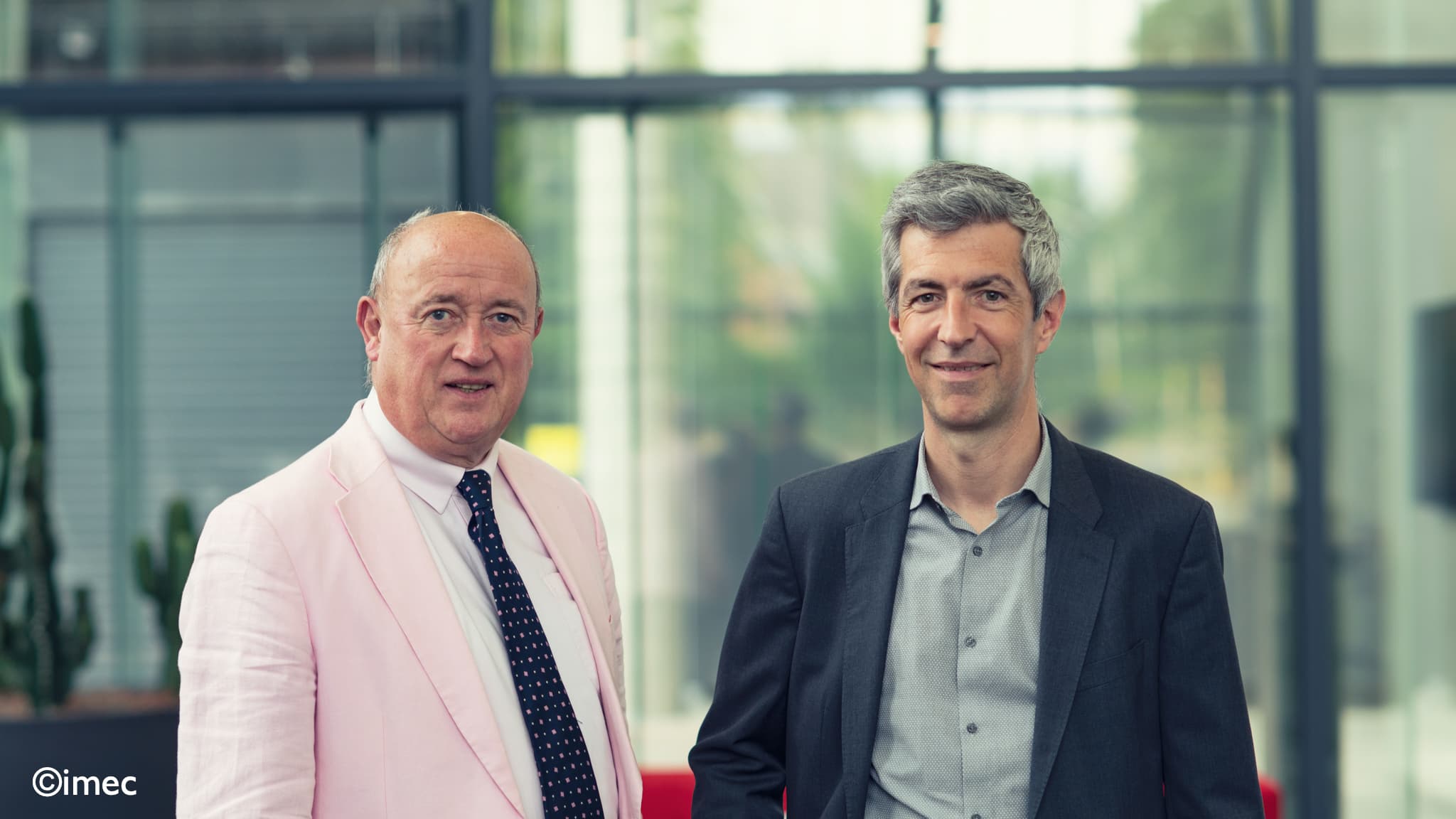 Huw-Hampson-Jones-%28Solithor%29-and--Olivier-Rousseaux-%28imec%29001-2 Two men in suits posing indoors in front of glass windows, one in a pale pink jacket and one in a dark blazer.