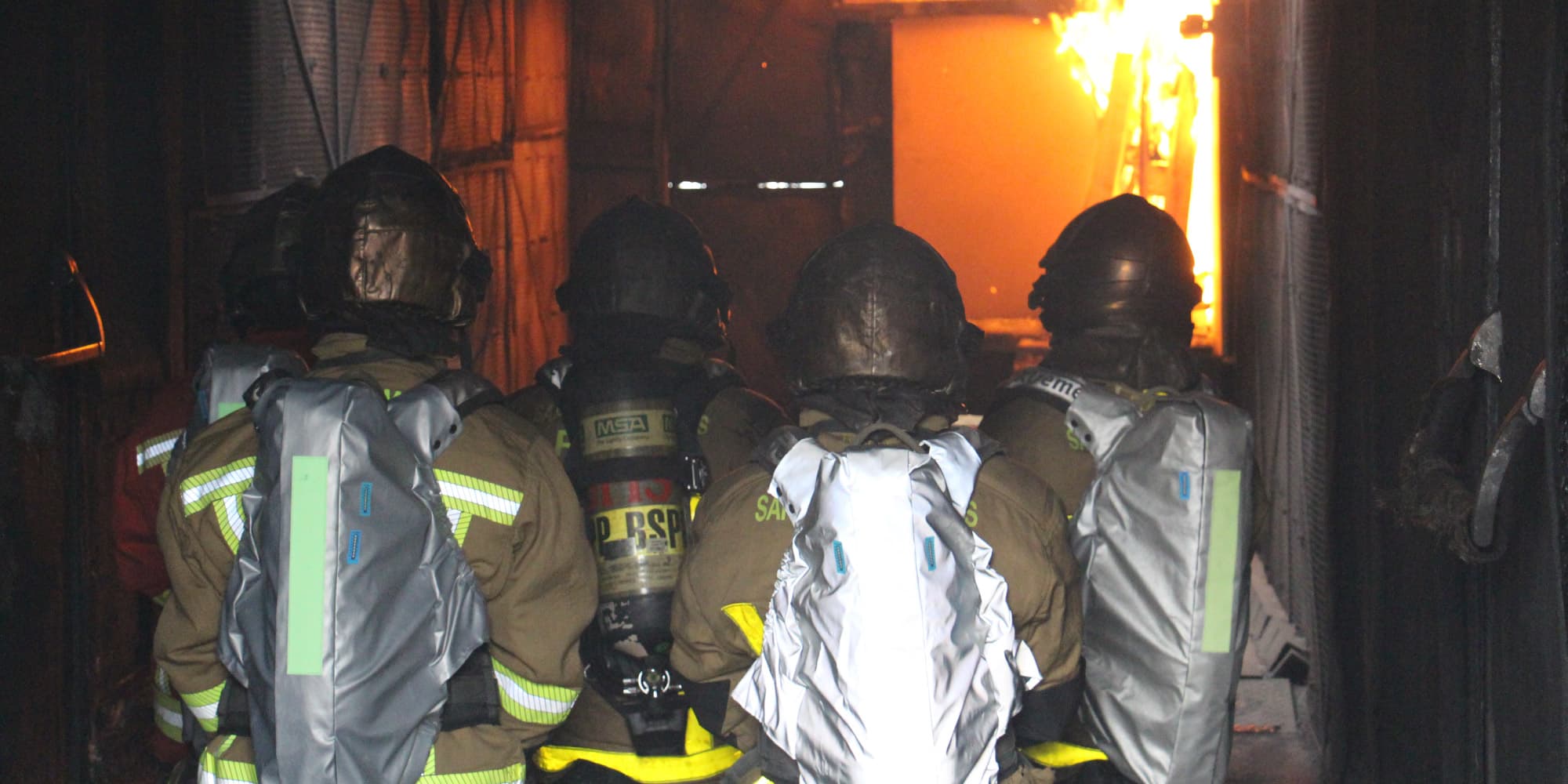 i-cart-press_1 Firefighters in protective gear move toward flames in a smoke-filled interior, with sensor-equipped backpacks on their suits.