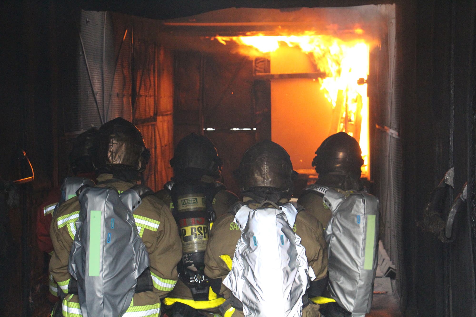 icart_brandweerpak_met_sensoren_5 Firefighters in protective suits advance through a burning corridor, with sensor-equipped packs visible on their backs.