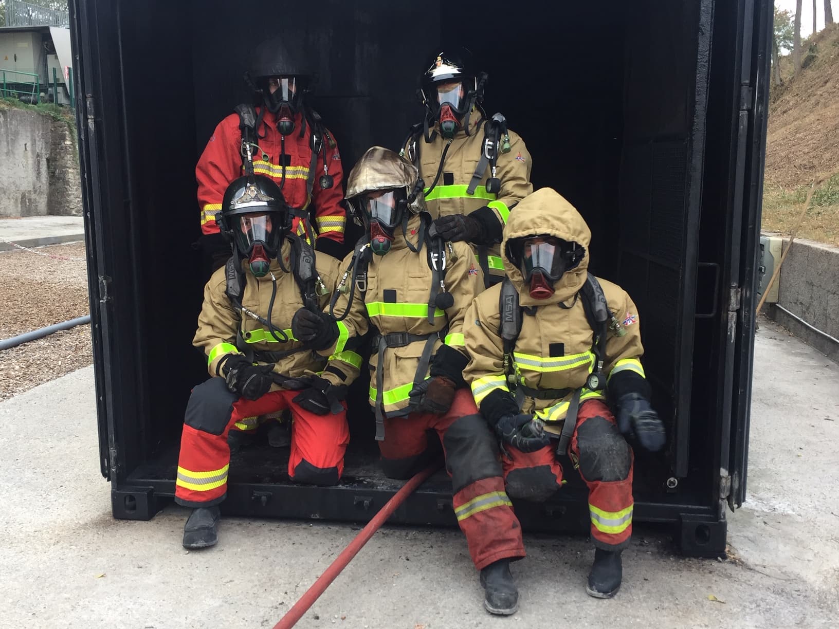 icart_brandweerpak_met_sensoren_6 Five firefighters in protective suits and breathing masks pose inside a dark training container