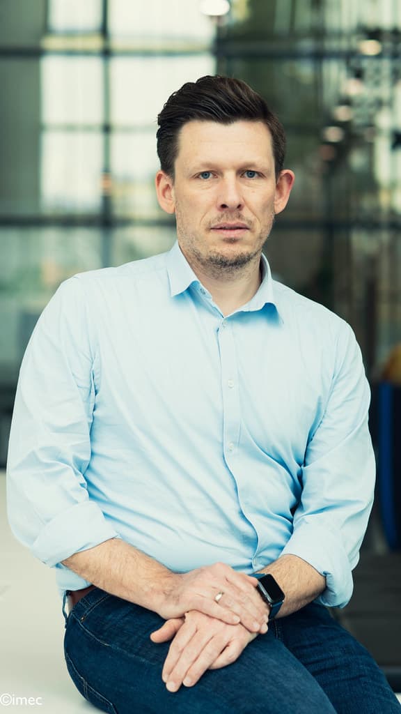 ilja-ocket Portrait of Ilja Ocket in a light blue shirt, seated with hands folded against a blurred glass background.
