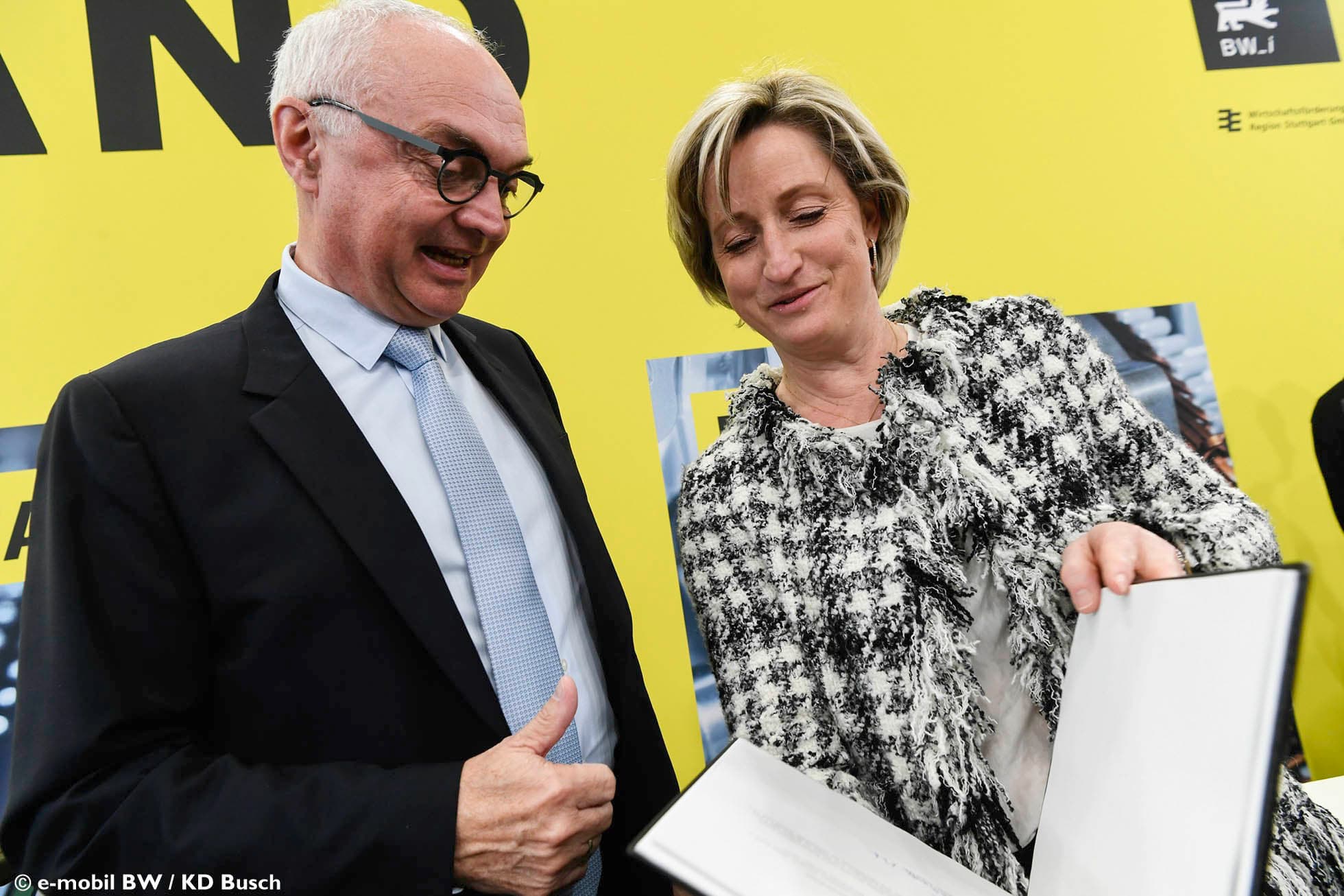 Two officials viewing presentation folder at imec Germany event Two officials smile while looking at an open presentation folder in front of a yellow imec Germany backdrop.