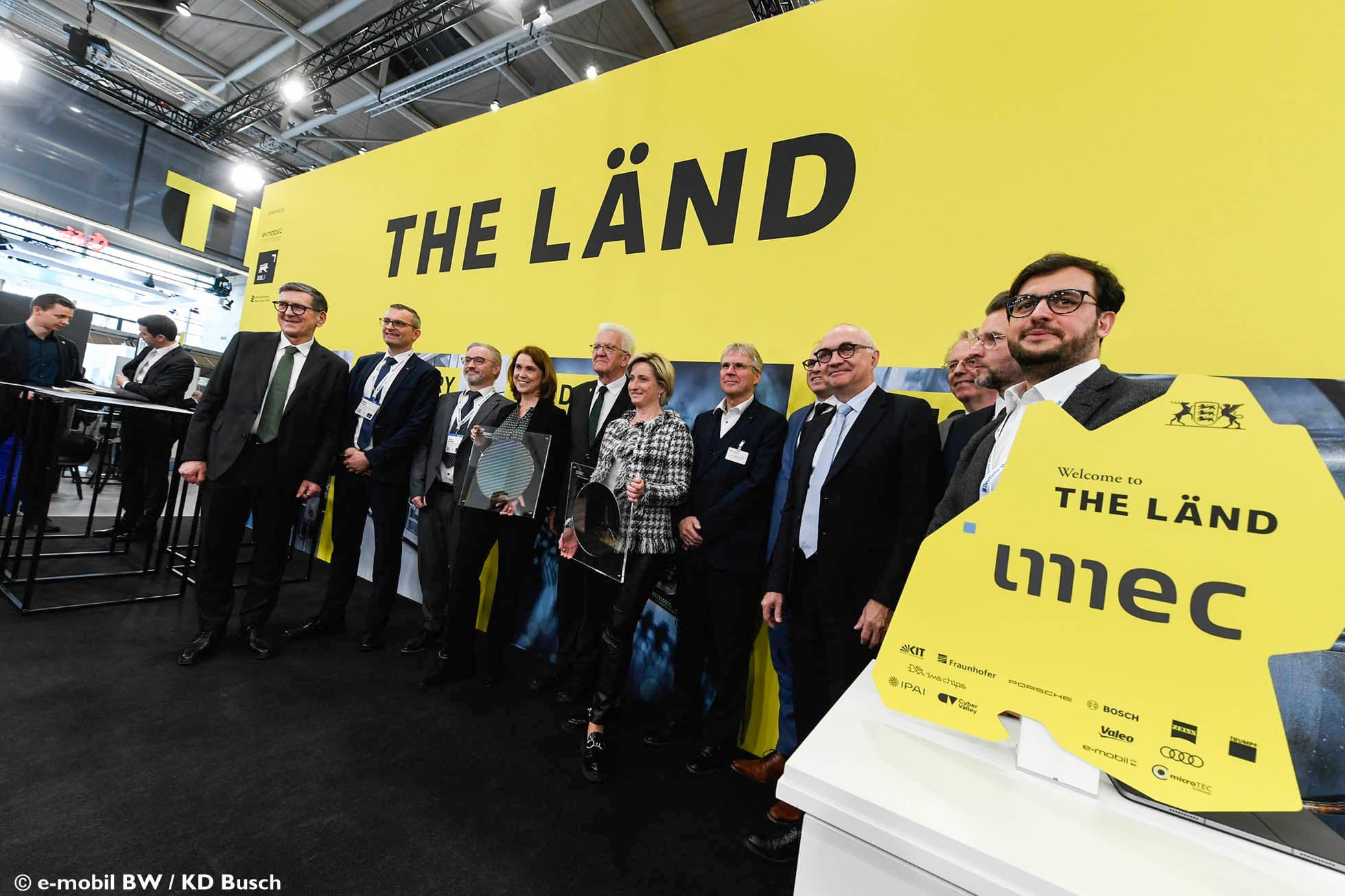 Officials posing at imec Germany booth with THE LÄND display Group of officials posing at imec Germany booth beside a large yellow THE LÄND display