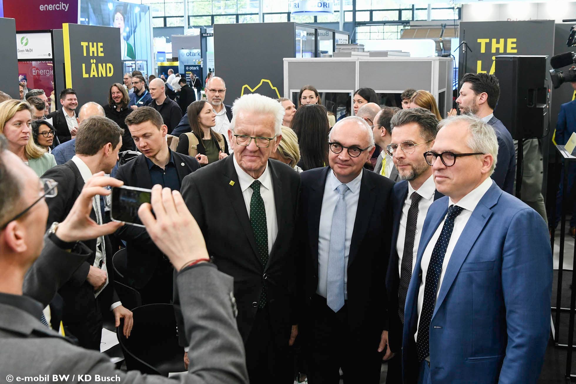 Group of officials posing at THE LÄND exhibition booth Groep in pak geklede functionarissen die poseren voor een foto bij een beursstand met "THE LÄND"-borden