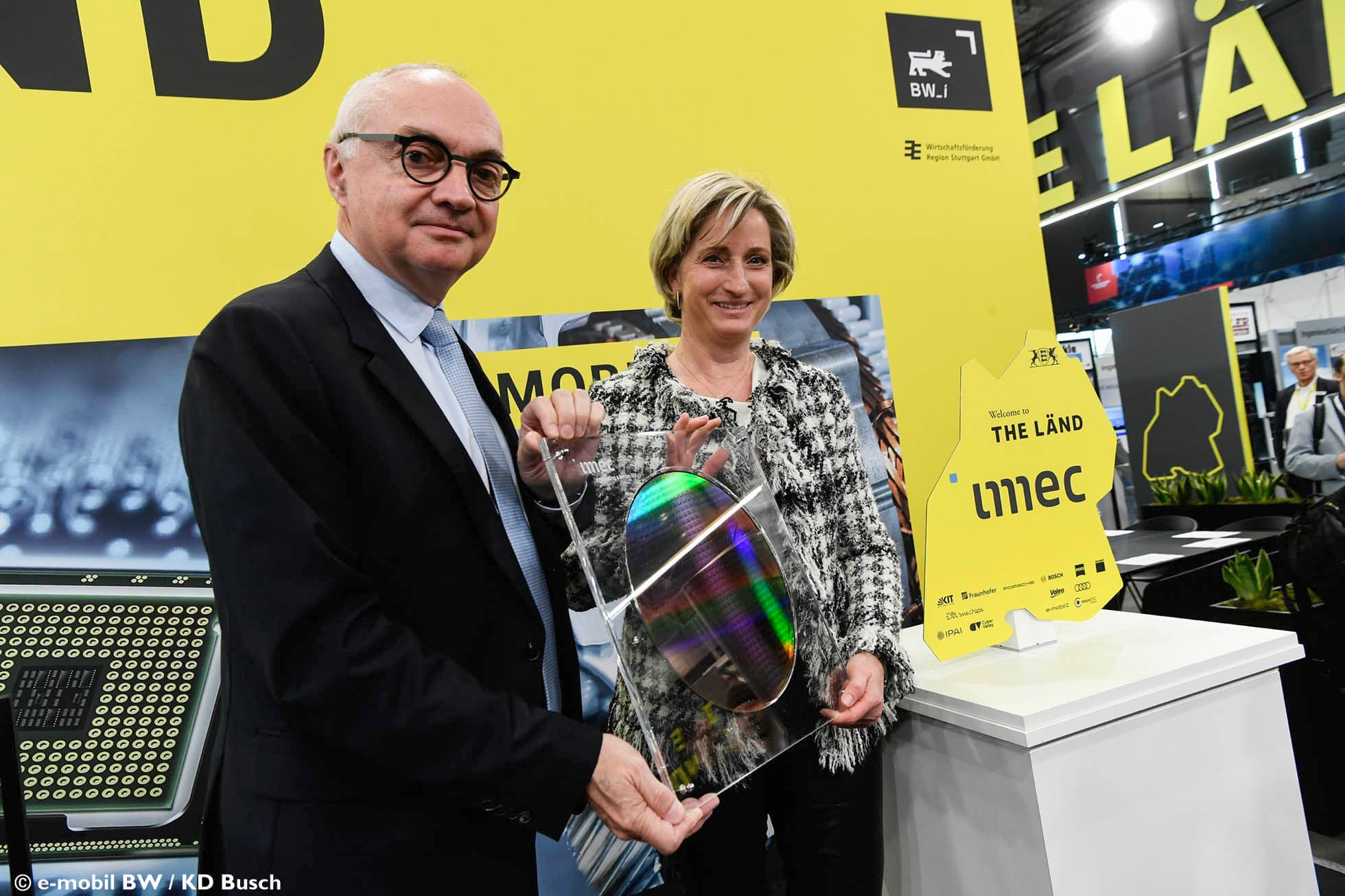 Officials holding semiconductor wafer display at imec Baden-Württemberg booth Two officials pose at a trade fair holding a semiconductor wafer display beside an imec Baden-Württemberg sign.