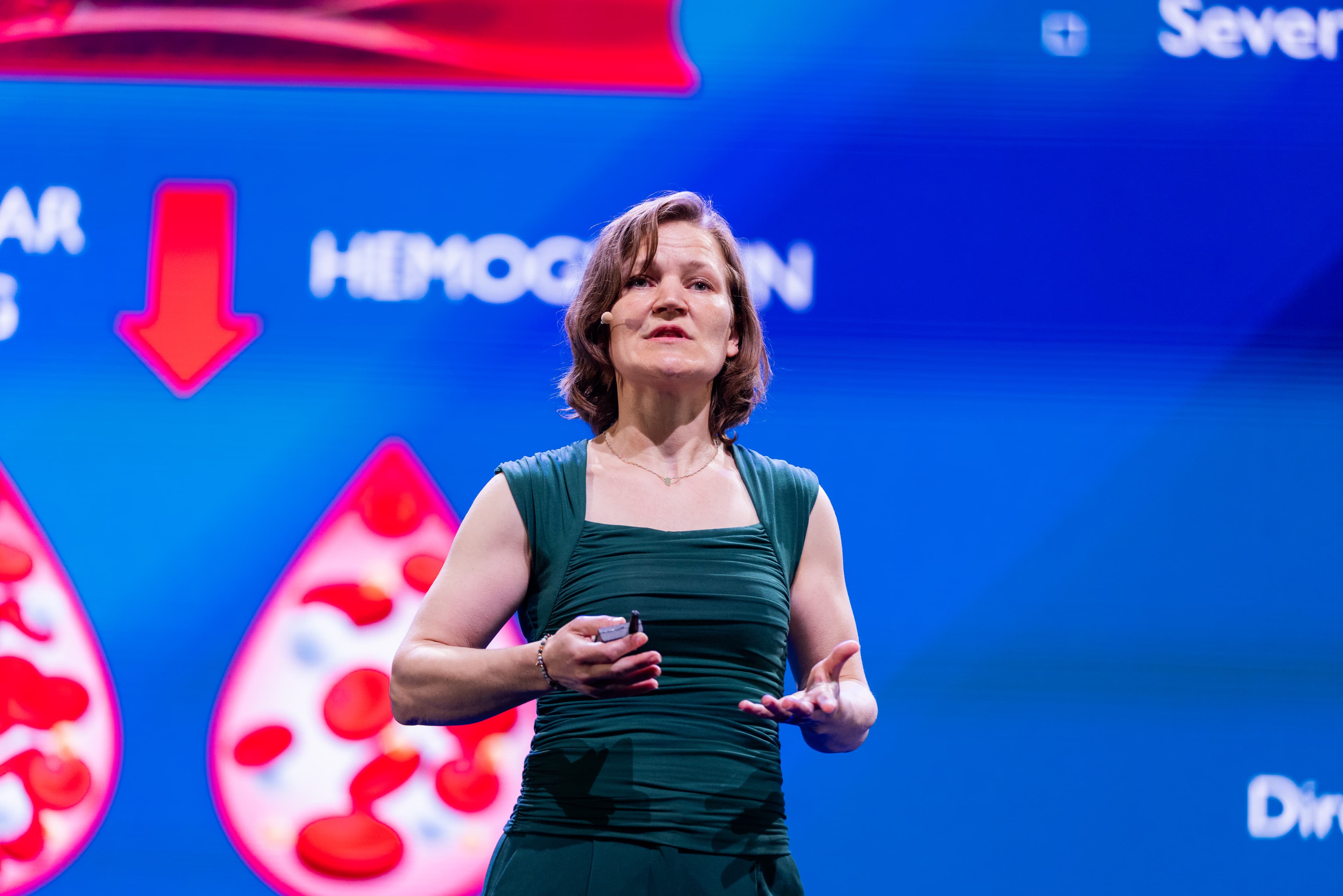 IMEC%20dag%202%20II_4 Speaker on stage presenting against a blue event backdrop with red arrow and cell graphics