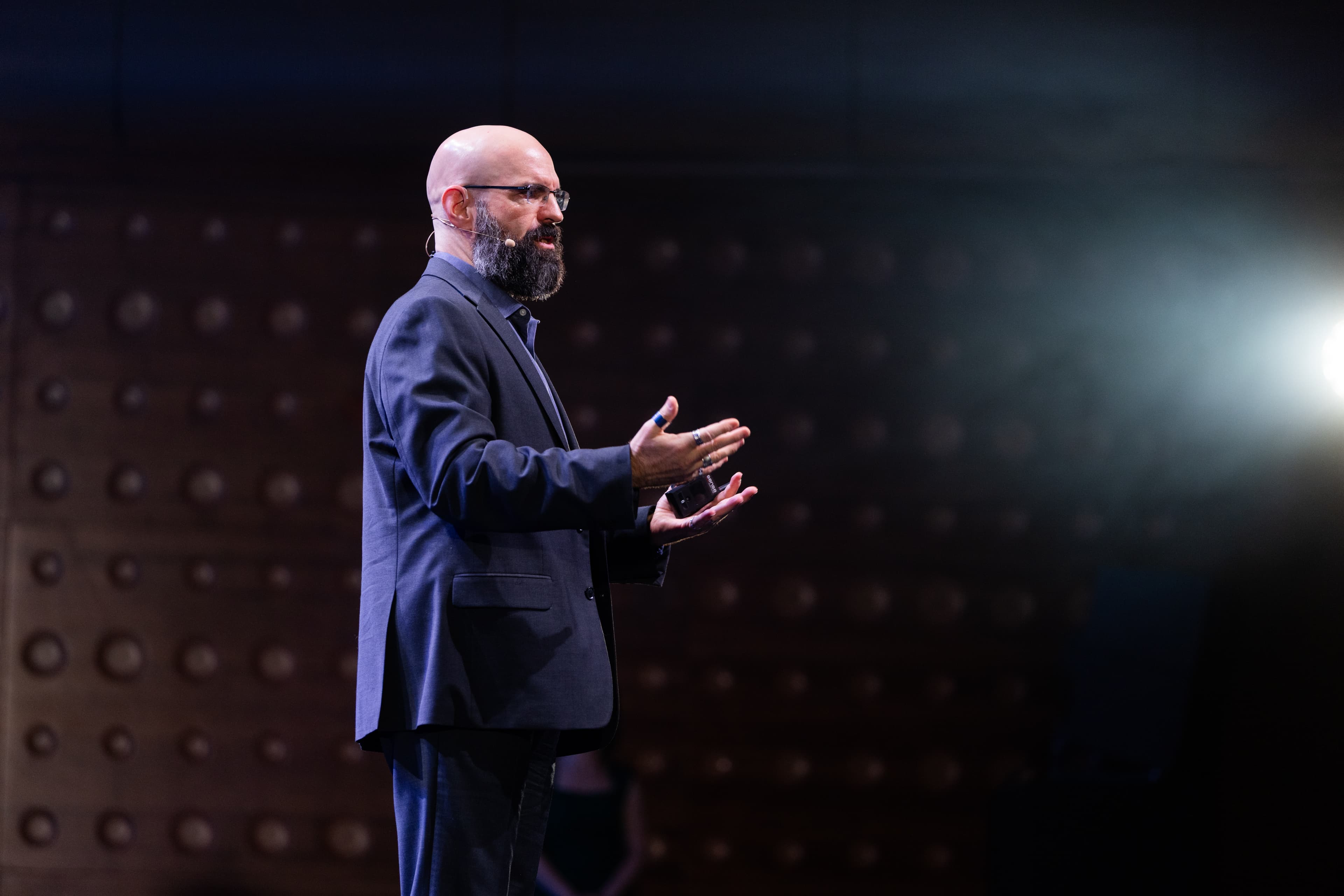 Speaker on stage with headset microphone A bald bearded speaker in a dark suit gestures on stage with a headset microphone.