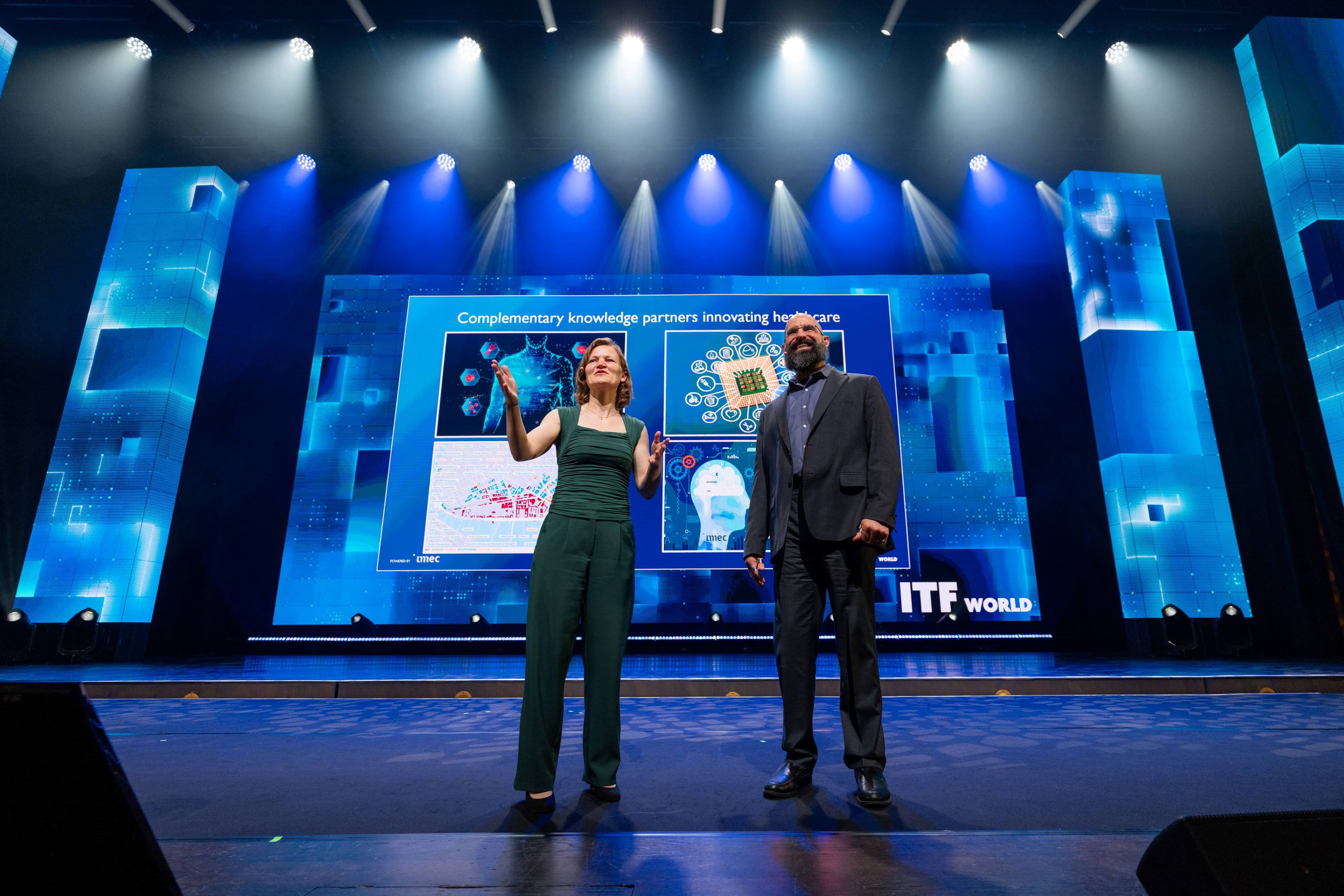 IMEC%20dag%202%20II_8 Two speakers on a blue-lit stage in front of a healthcare presentation screen