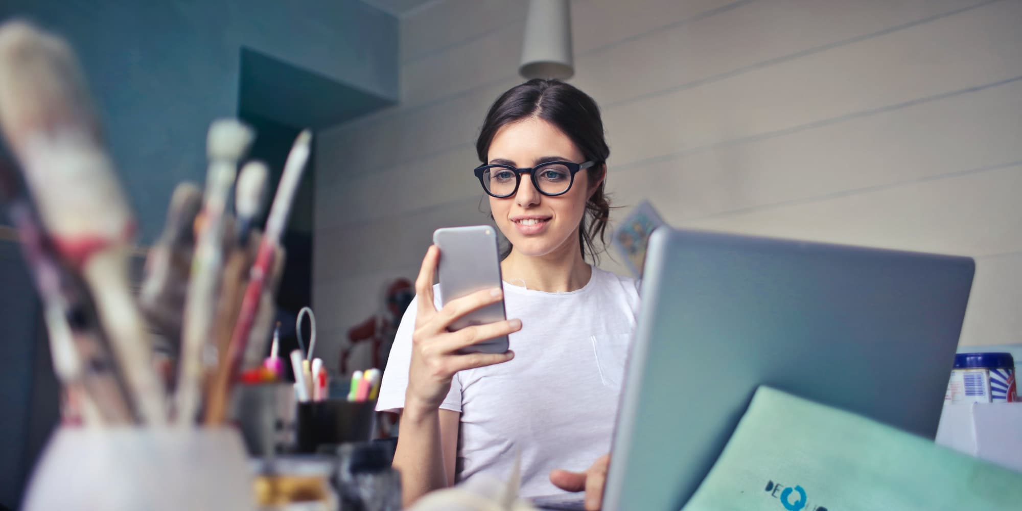 imec.digimeter-2022 Jonge vrouw die haar smartphone bekijkt naast een laptop op een bureau