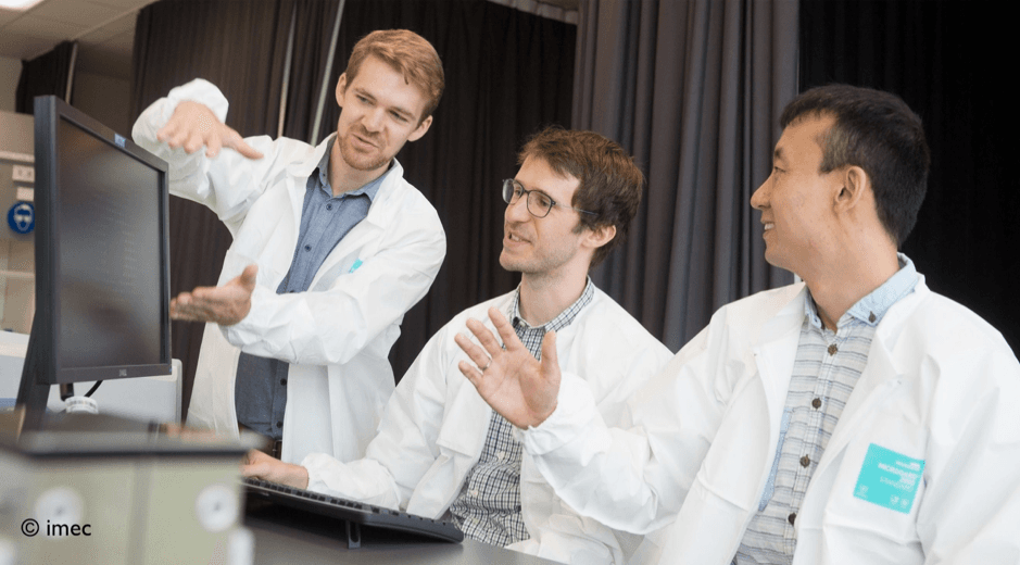imec_erc_grant_1 Three researchers in lab coats discuss a computer screen in a laboratory setting.