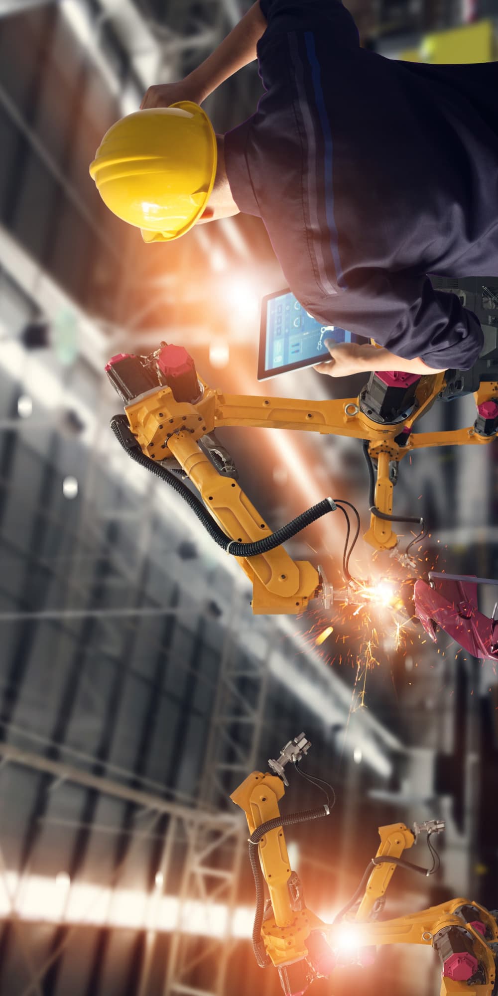 imec-magazine-ai-special-07_pocs_humanai_1 Worker in a yellow hard hat controls robotic arms welding in an industrial setting.