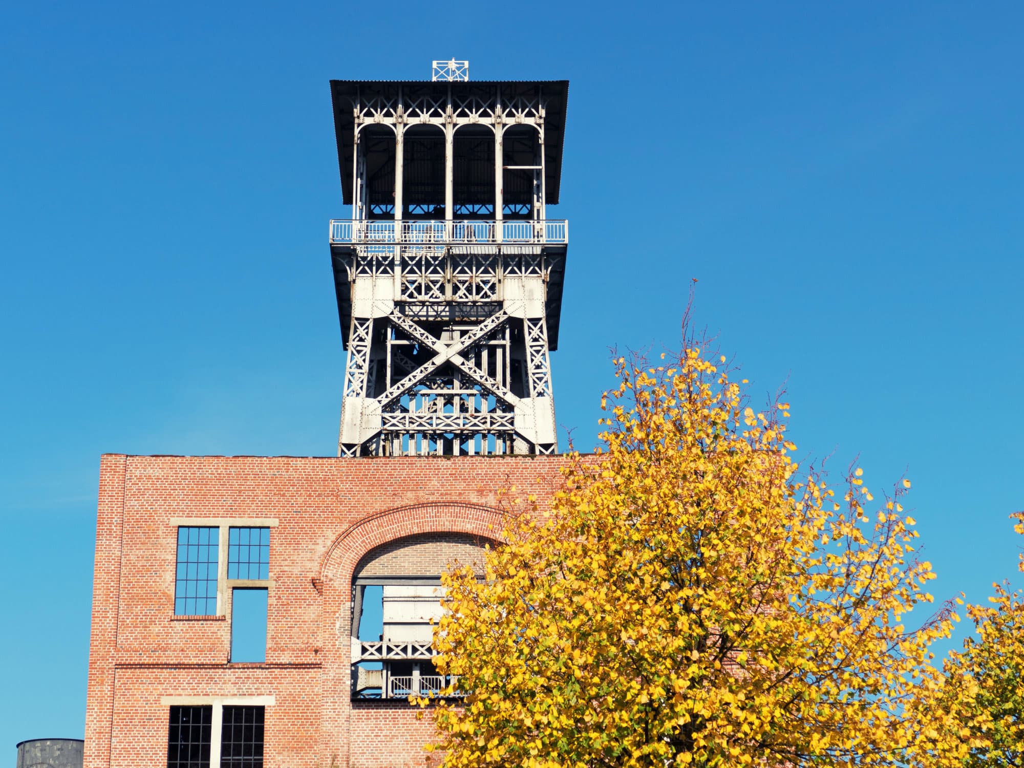imec-magazine-genk-energyville_1 Brick industrial building with a steel tower rising above autumn trees under a clear blue sky