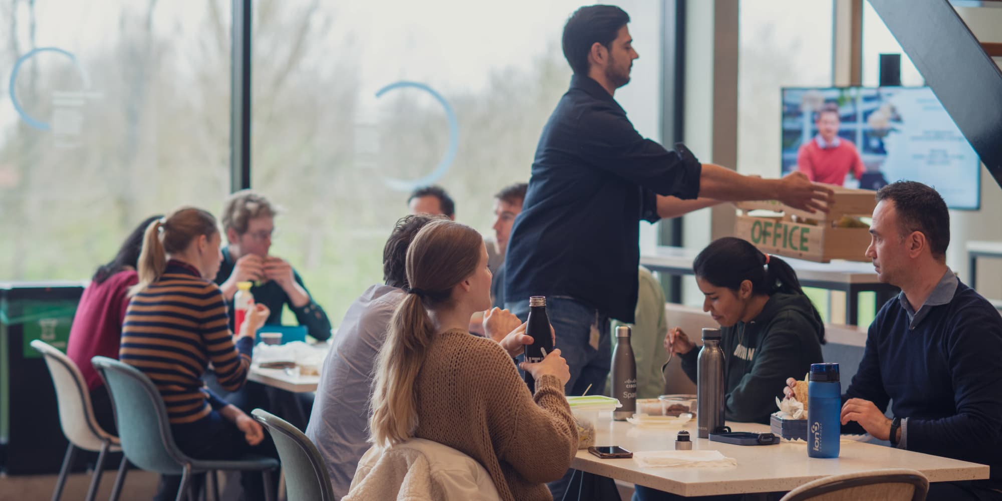 imec-the-Netherlands-benefits Colleagues sharing lunch at a cafeteria table while one person carries a box past them
