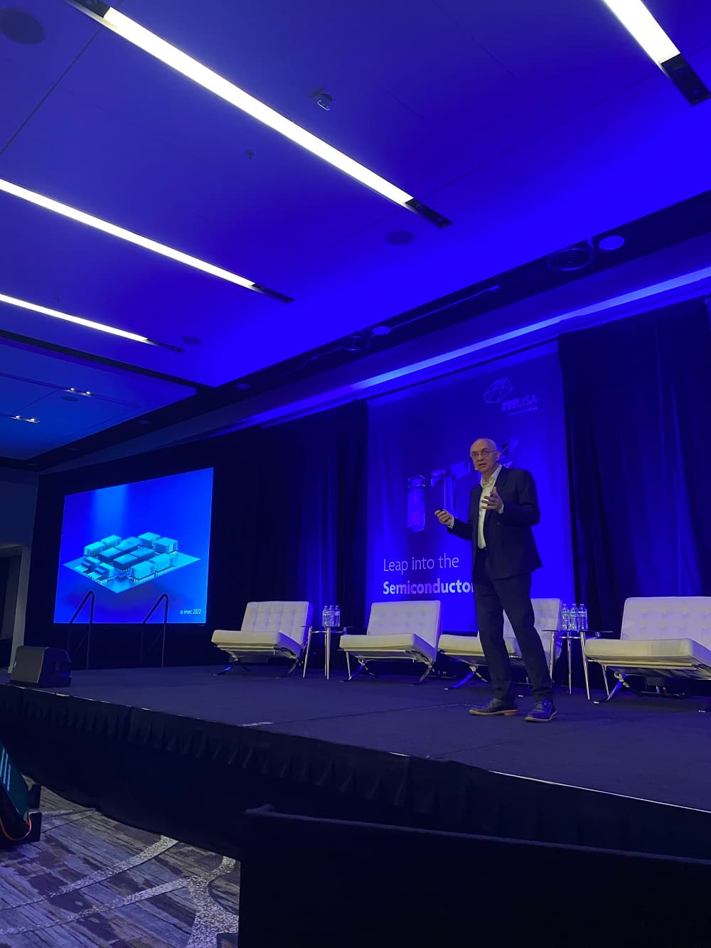 Speaker presenting on conference stage with chip slide Speaker presenting on a blue-lit conference stage beside a slide with a chip illustration.