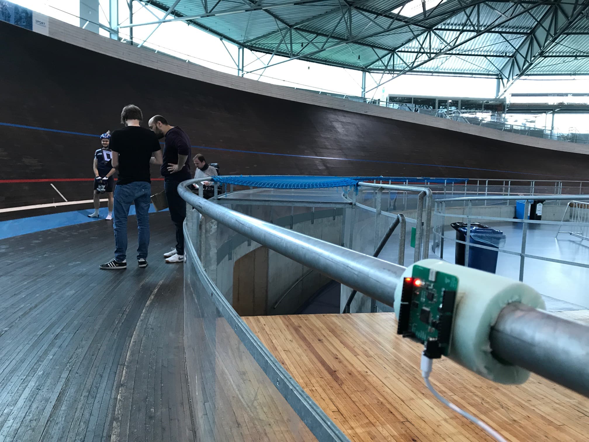 IoT%2520baanwielrennen%2520%25286%2529 IoT-sensor bevestigd aan een baanrail in een wielerbaan, met fietsers en onderzoekers op de achtergrond