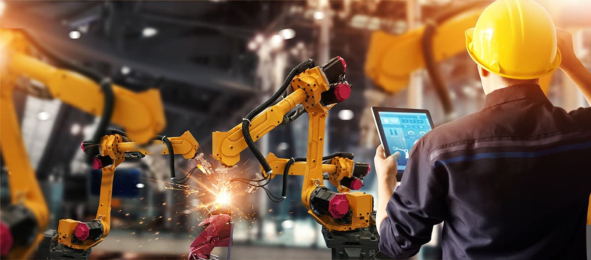 Worker monitoring robotic welding arms in factory Worker in a hard hat monitors industrial robot arms welding on a factory line