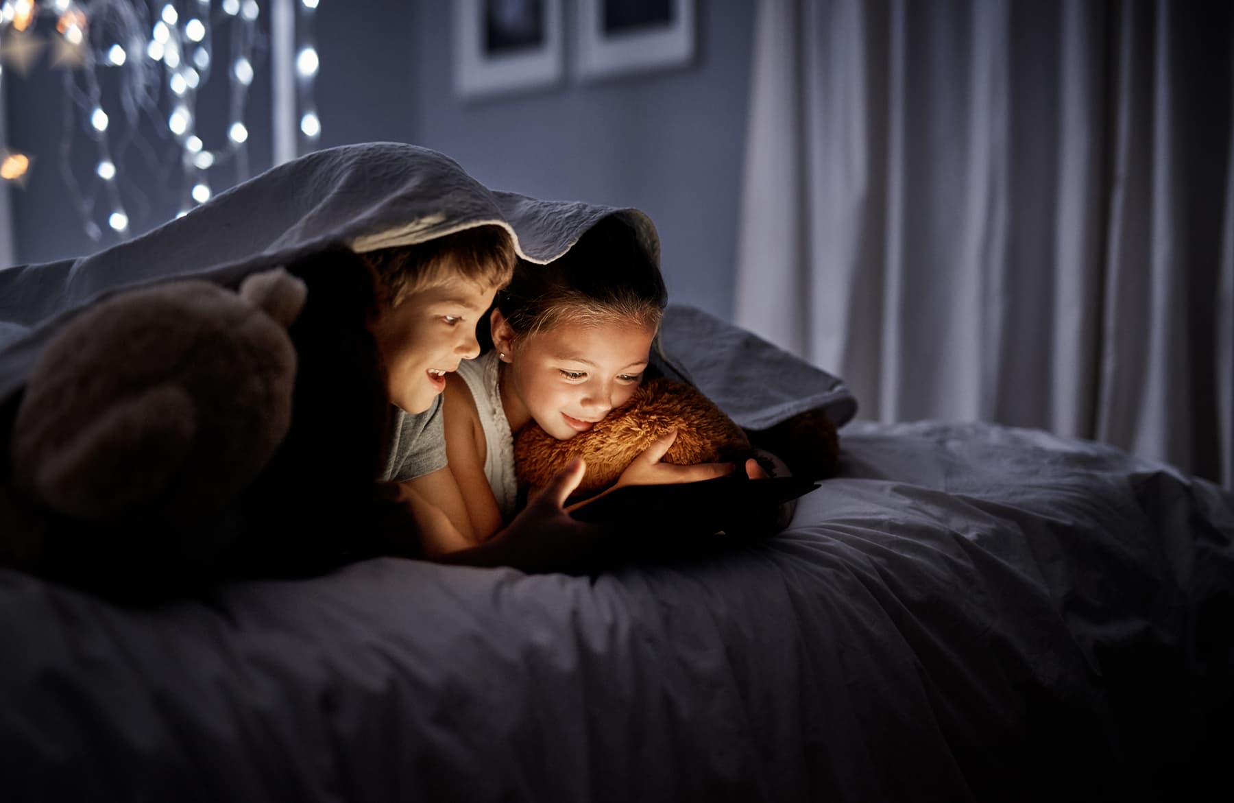 istock-1034734308_1 Two children under a blanket, looking at a tablet in a dimly lit bedroom
