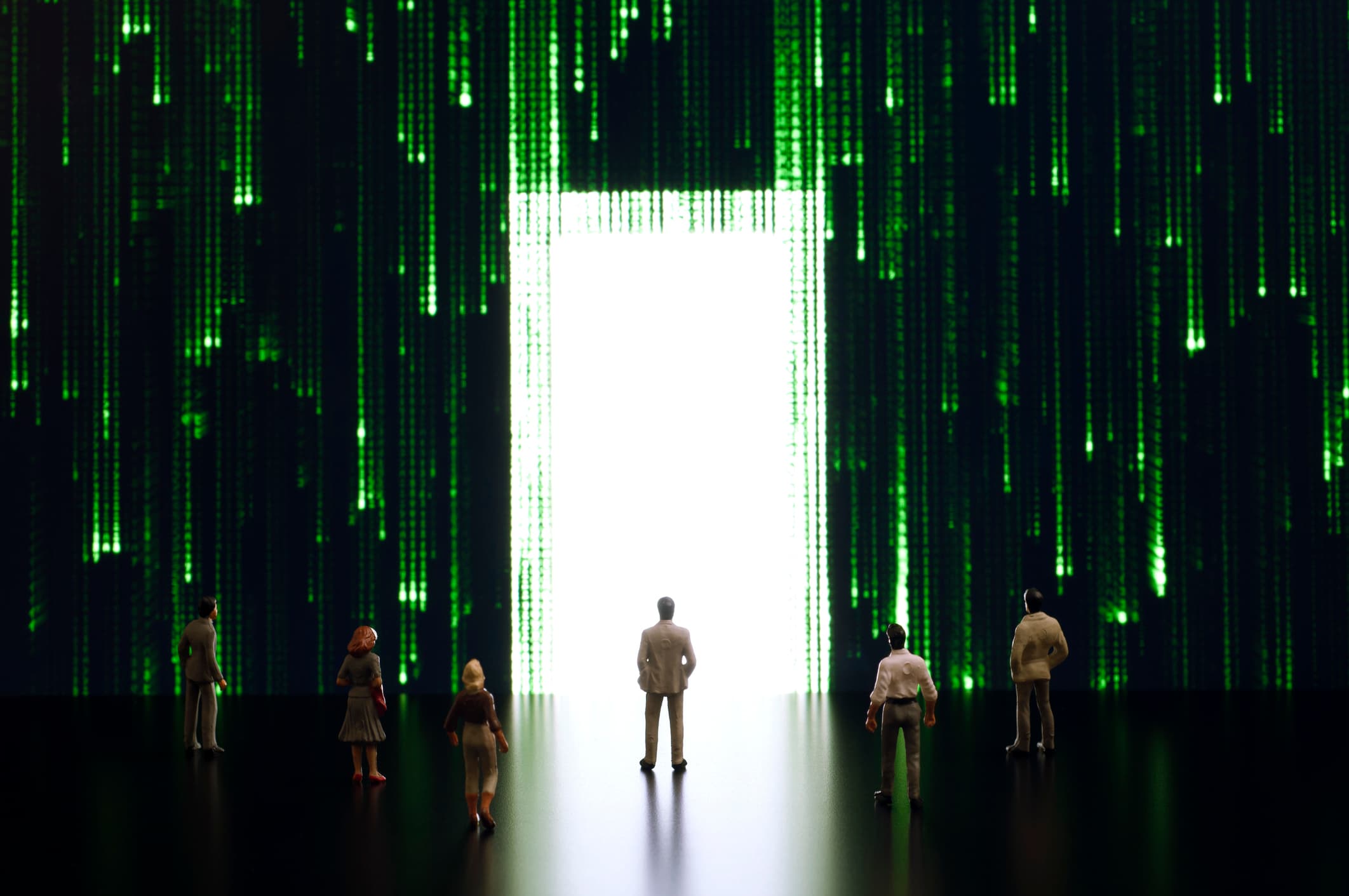 iStock-1329418153 Miniatuurmensen staan voor een gloeiende deuropening te midden van groene digitale code, wat een virtuele wereld oproept.