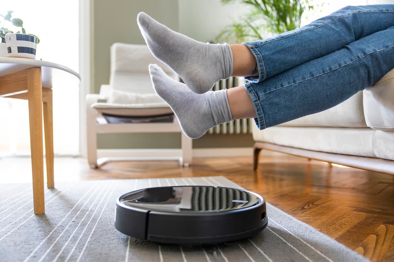 iStock-1331271160 Robotstofzuiger op een vloerkleed in de woonkamer naast iemands met sokken bedekte voeten op een bank