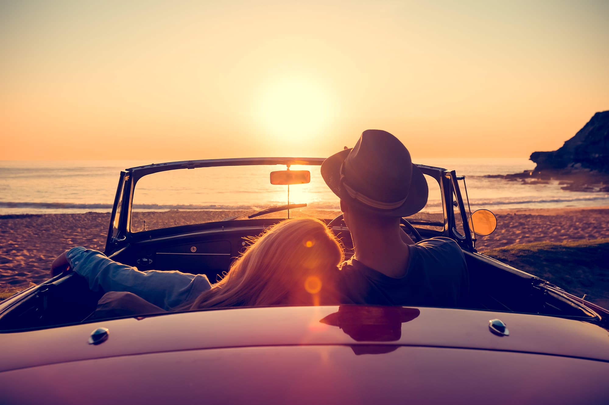 iStock-514059558_1 Couple sitting in a convertible by the sea at sunset, looking toward the horizon