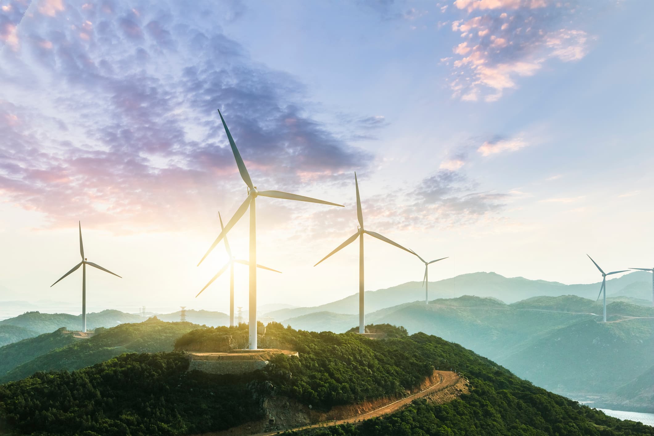 Wind turbines on mountain ridge at sunrise Wind turbines on a green mountain ridge at sunrise, with a winding dirt road and pastel clouds overhead.