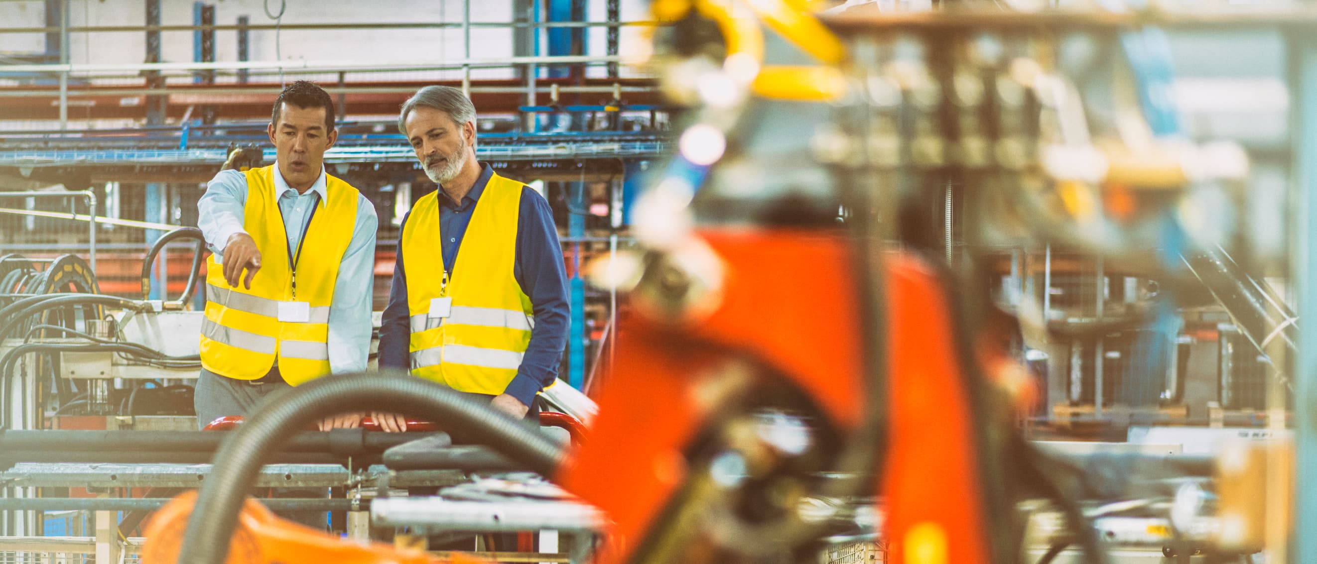 iStock-892766028 Two workers in yellow safety vests discuss a factory floor beside blurred machinery and equipment.