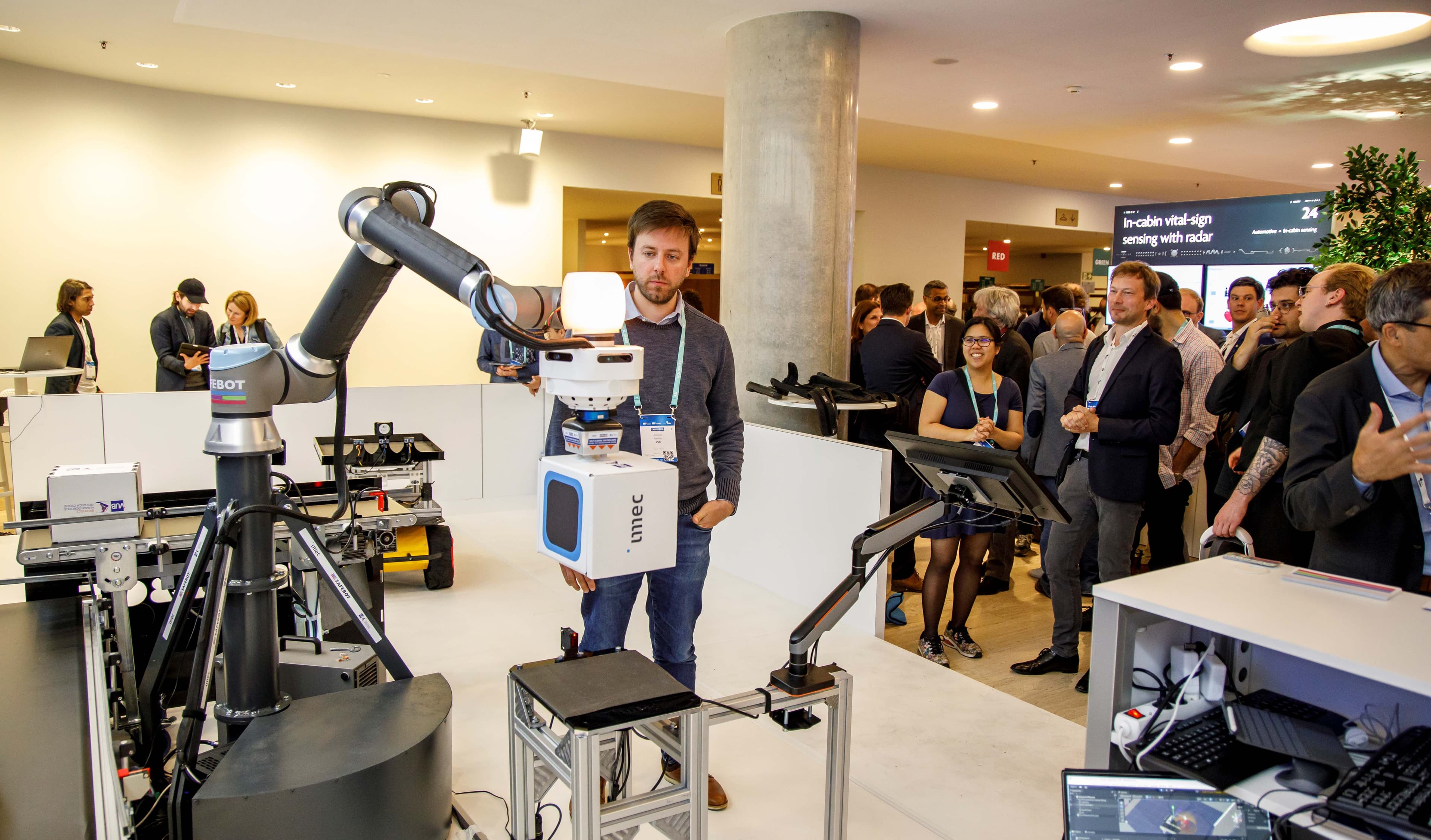 Demonstration eines Roboterarms auf einer Messe mit Publikum A demonstration robot arm beside a man at a crowded event booth, with attendees watching sensor equipment