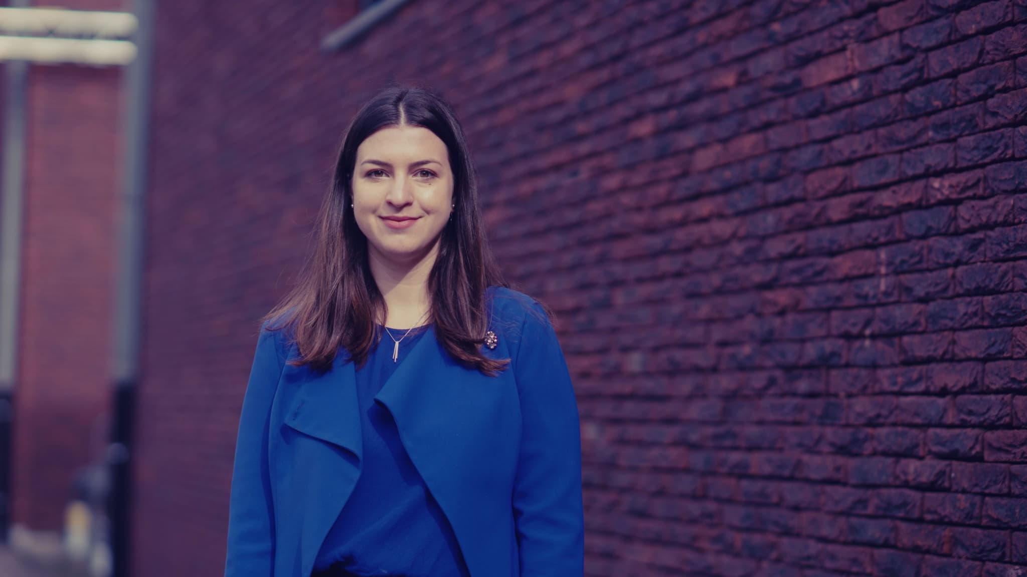 Iva%20Popova00000_maxWidth_2048_maxHeight_1152_ppi_72_quality_100_embedMetadata_true_contentETag_1 Portrait of a woman in a blue jacket standing against a brick wall