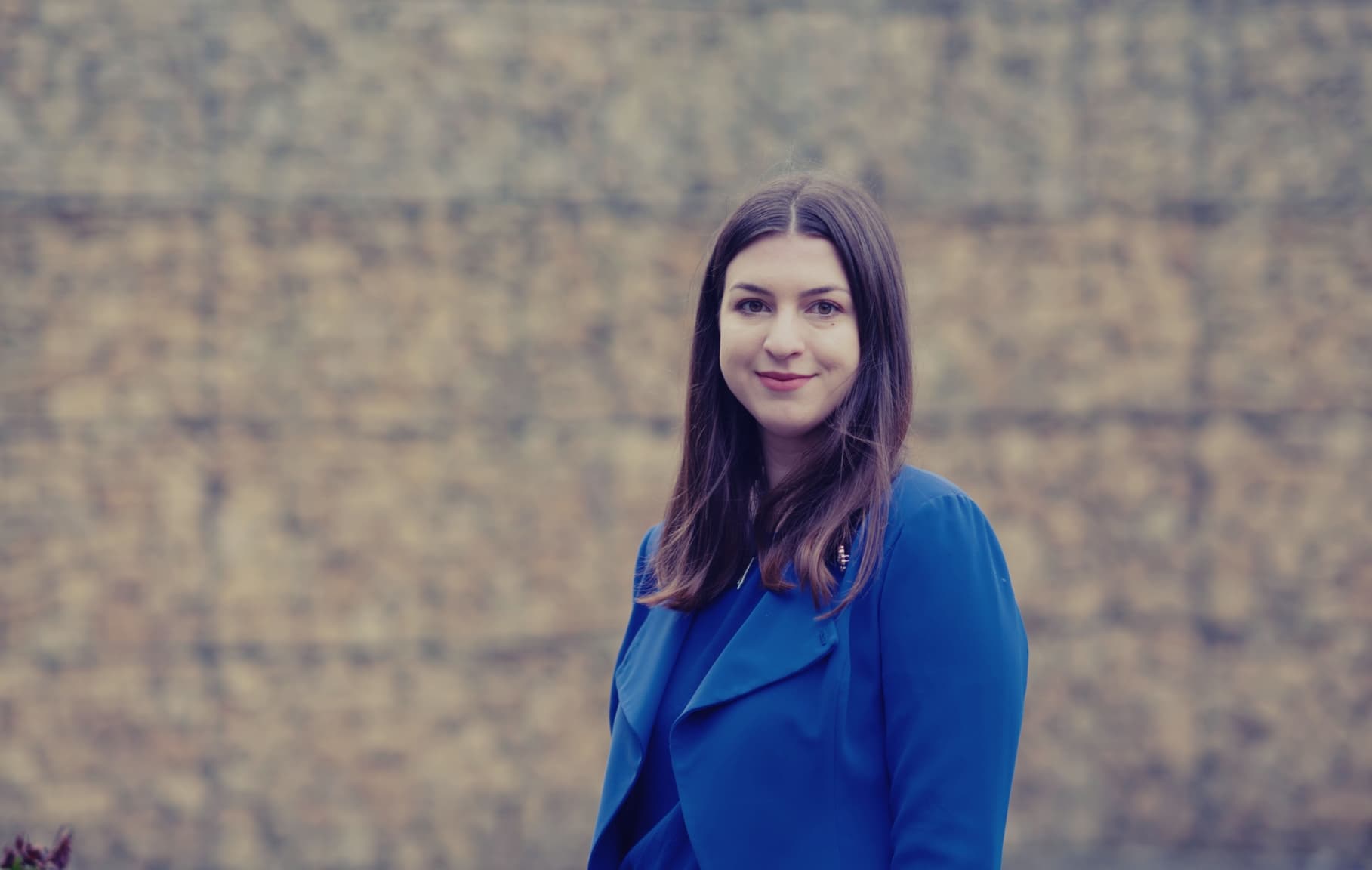 Iva%20Popova00003_maxWidth_2048_maxHeight_1152_ppi_72_quality_100_embedMetadata_true_contentETag_1 Portrait of a young woman in a blue jacket standing against a blurred stone wall.