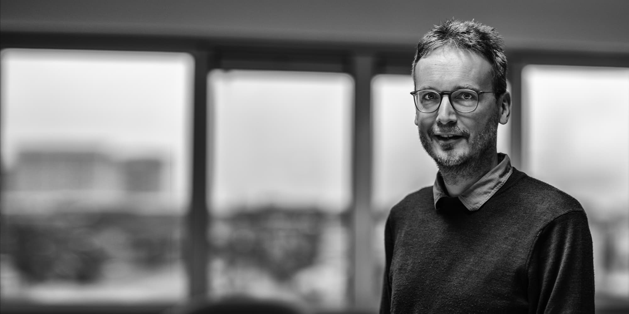 J.Adriaansen001magazinenew_1 Zwart-witportret van een man met een bril, staand voor een wazige achtergrond van een raam.