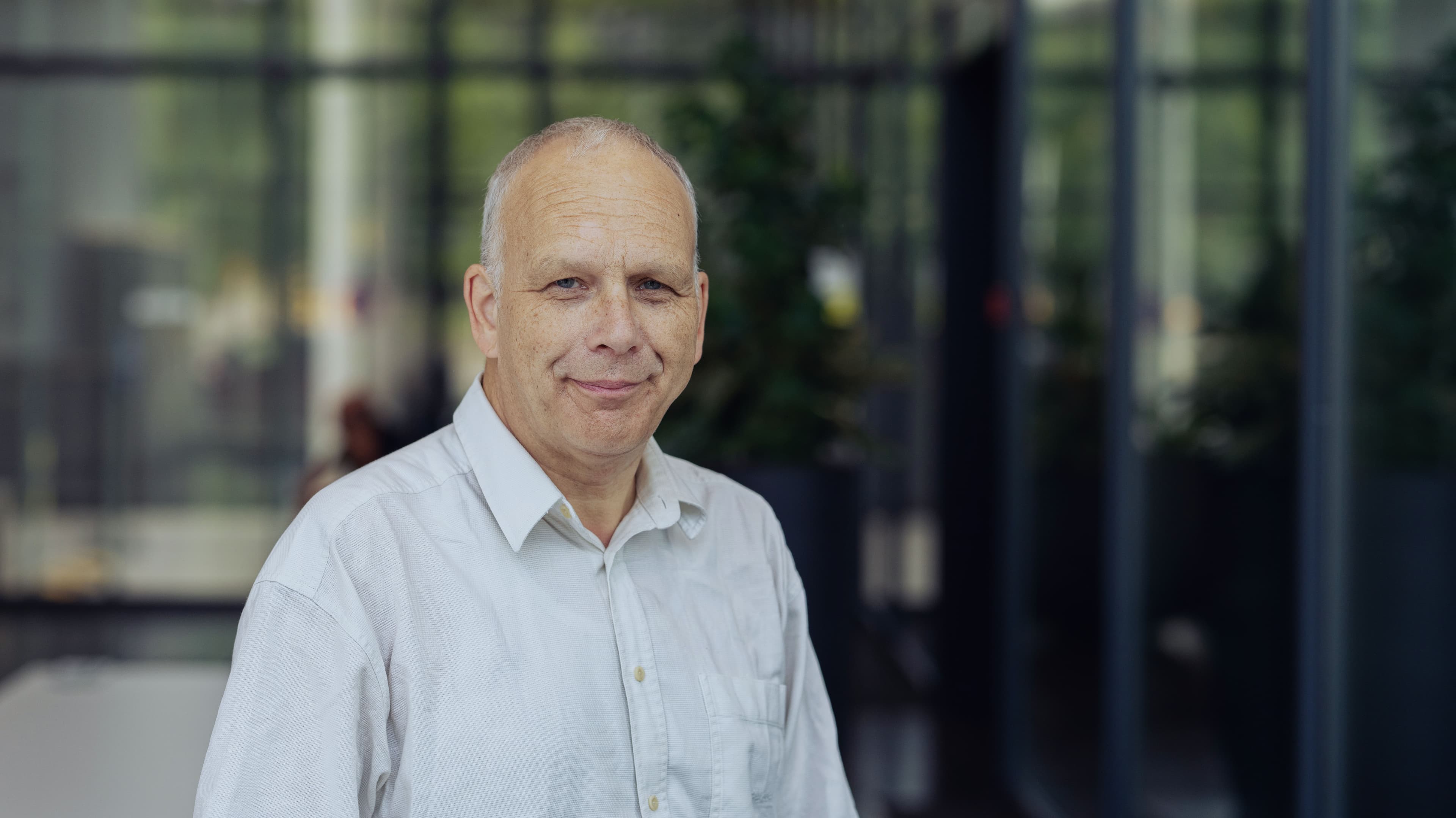 Jan%20Genoe001 Portrait of imec fellow Jan Genoe in a white shirt, standing indoors against a blurred glass background.