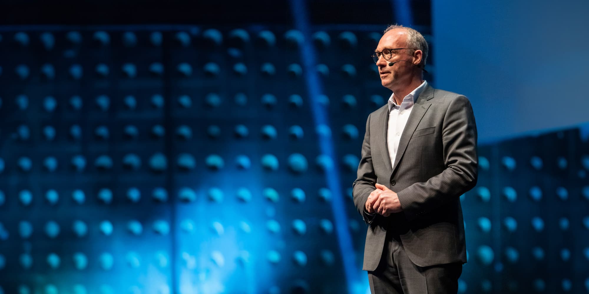Jo-De-Boeck-ITF Man in a gray suit speaking onstage with a headset microphone against a blue-lit backdrop