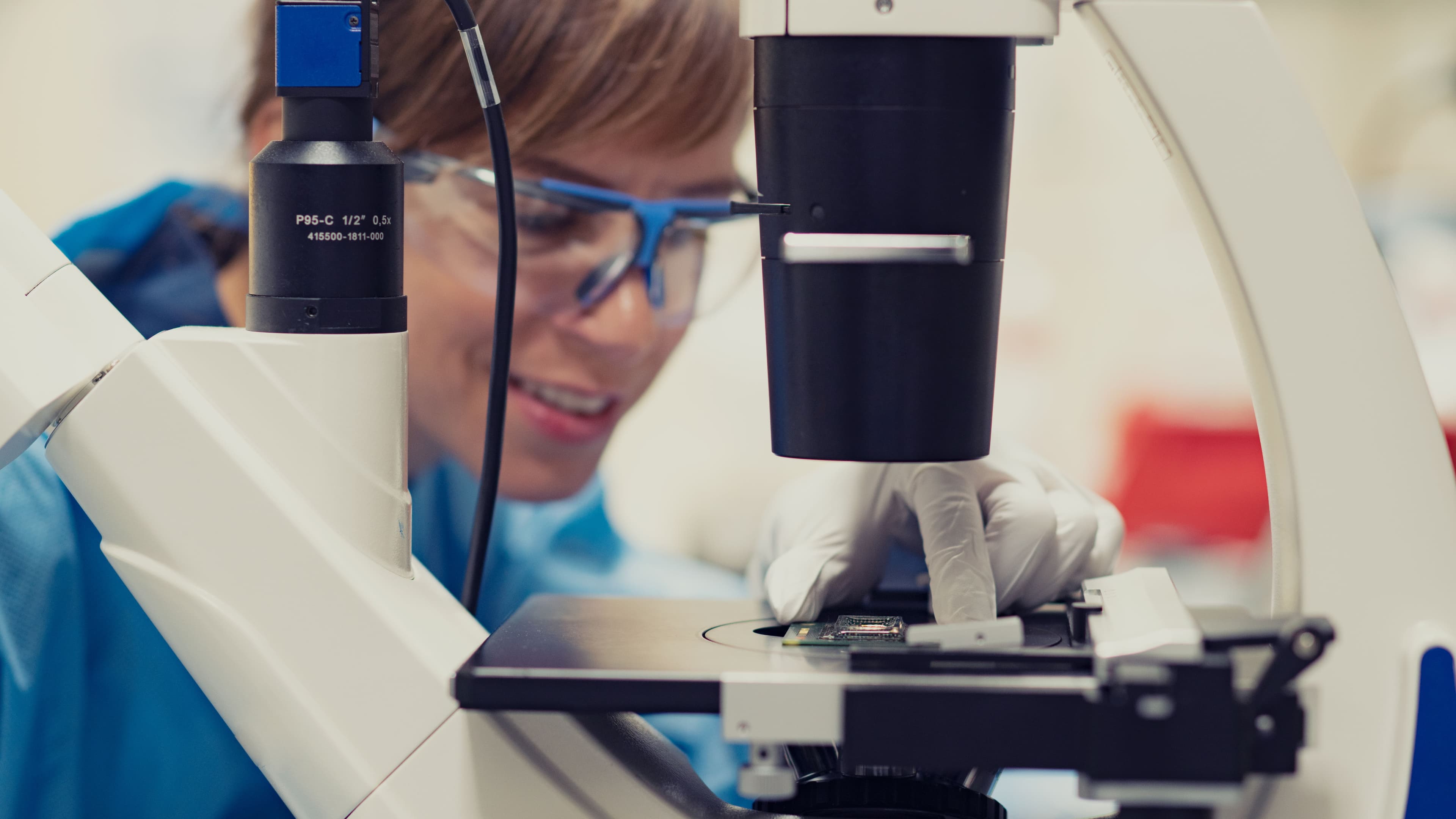 Johanna%20micro Researcher in a lab coat and gloves adjusting a microscope to inspect a sample on the stage.