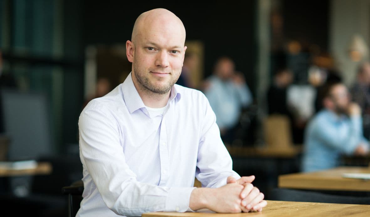 Joris_Van_Campenhout_LR_1 Joris Van Campenhout zit aan een tafel in een wit shirt en poseert voor een portret in een onscherpe kantooromgeving.