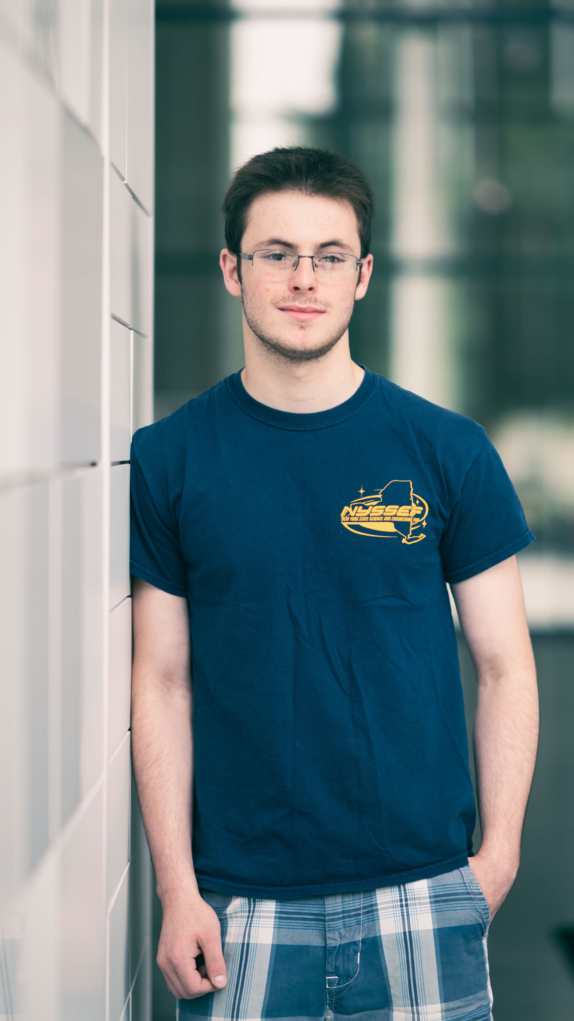 Katrien-mols006_Ariel_1 Young man in glasses wearing a NYSSEF T-shirt, standing indoors and facing the camera.