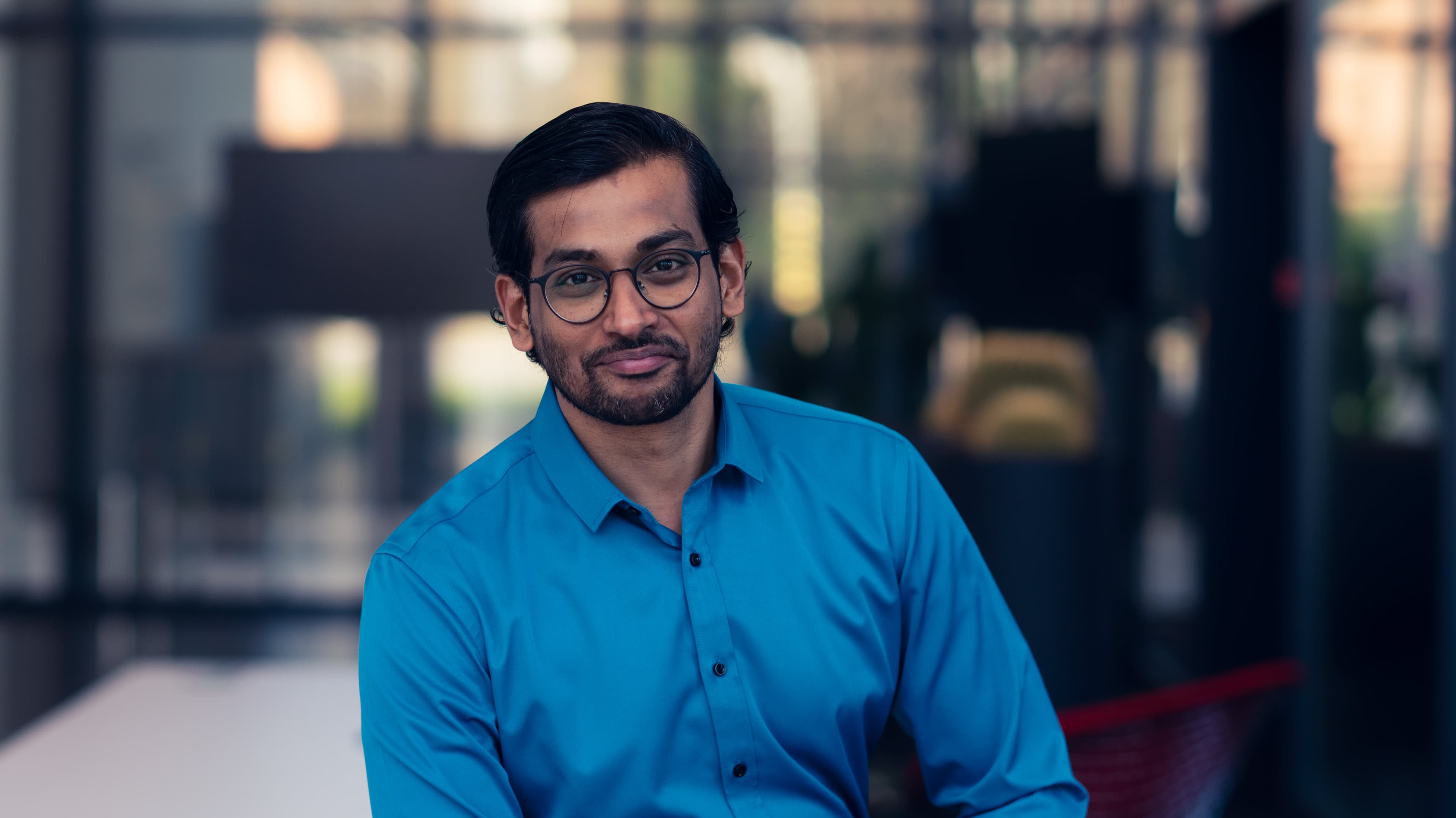 Kaustuv-Banerjee001-min Portrait of a man in a blue shirt and glasses standing in front of a blurred office background.