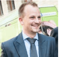 koen_triangle02_1 Smiling man in a dark suit and tie, shown in a head-and-shoulders portrait