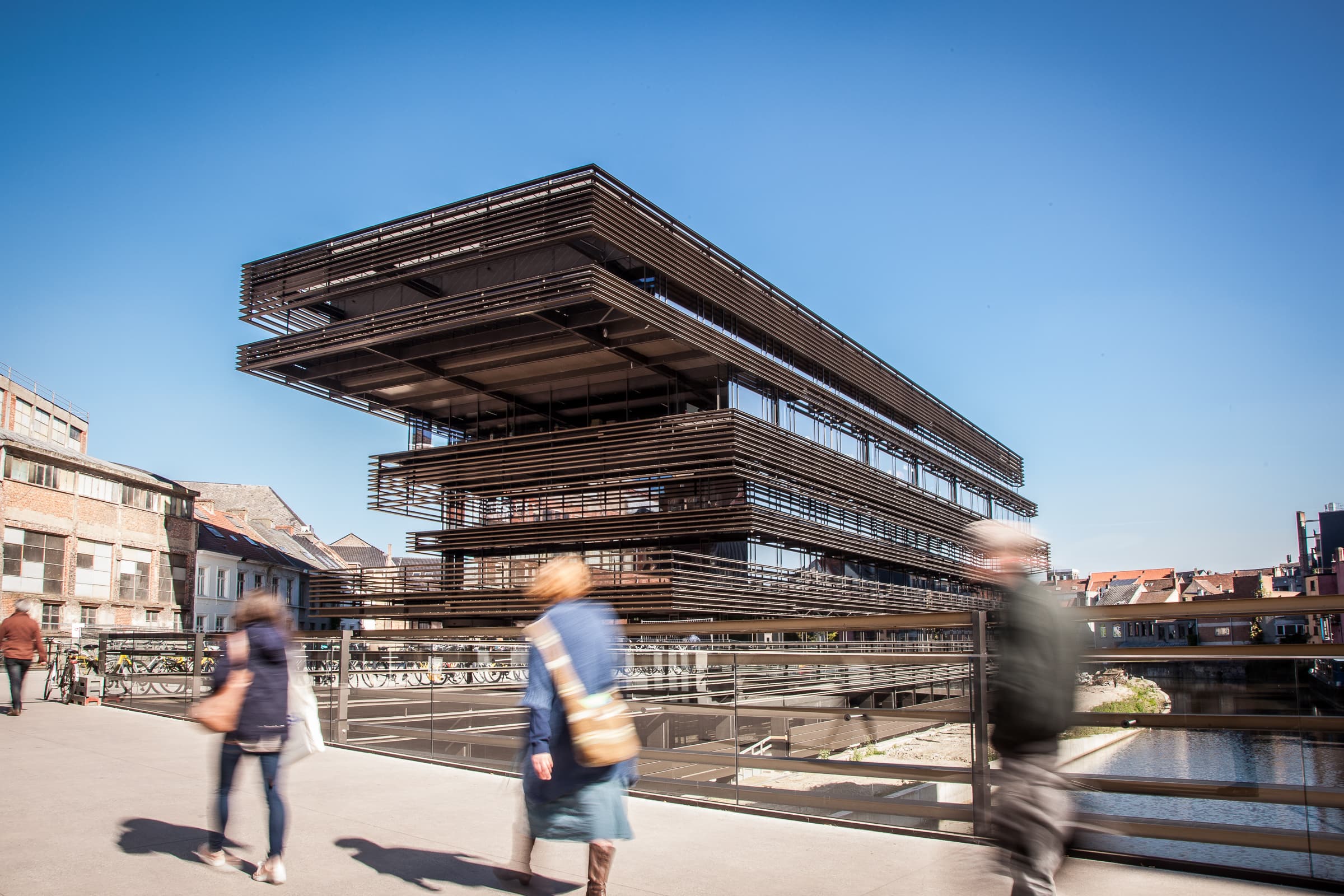 Krook2_0 Modern riverside public building with people walking in the foreground under a clear blue sky.