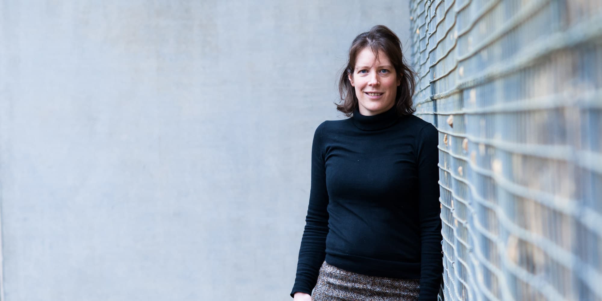 liesbet-lagae_1 Portrait of a woman standing beside a wire fence, wearing a black turtleneck and patterned skirt
