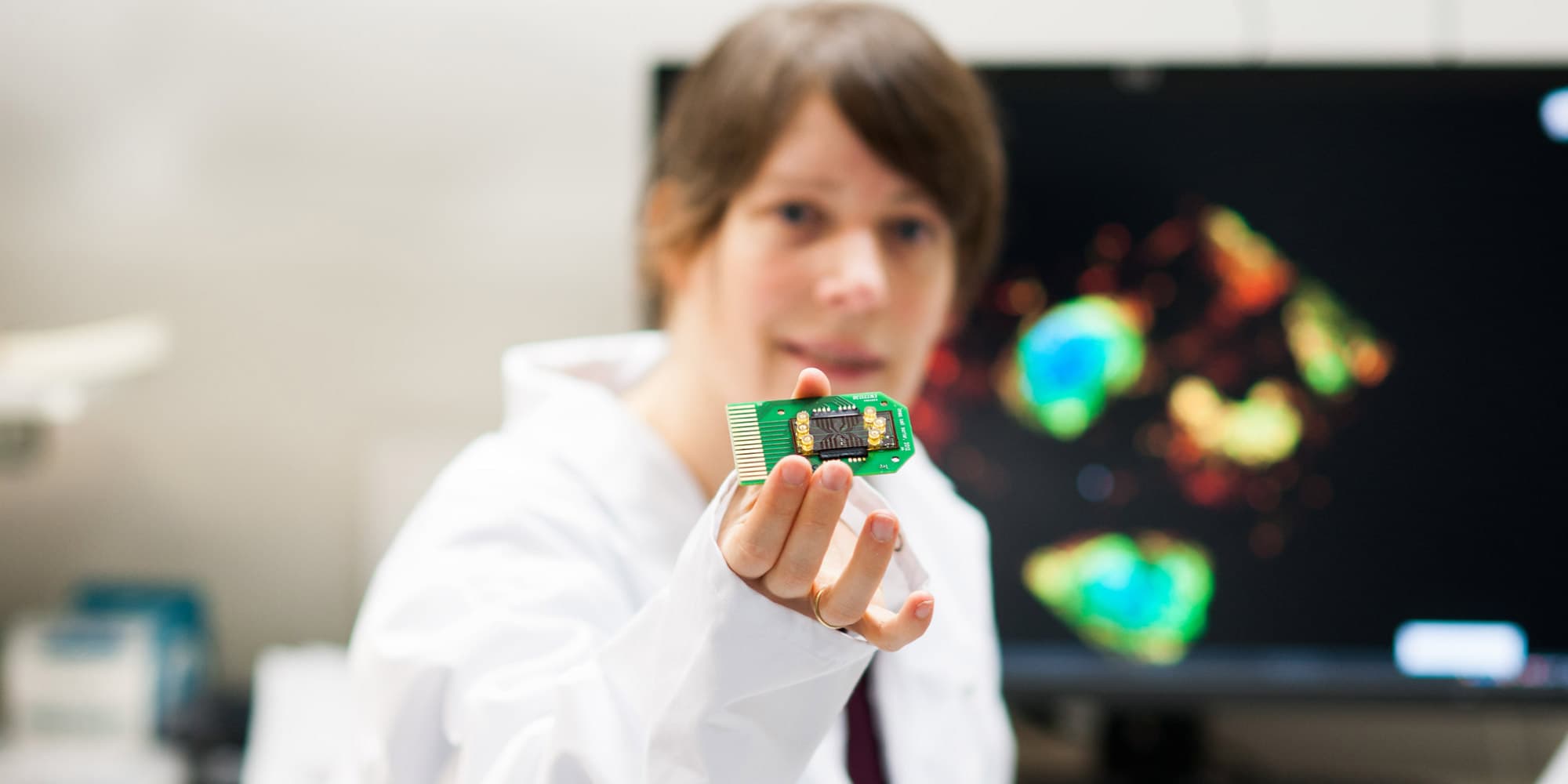 Liesbet-Lagae Researcher in a lab coat holding up a small green circuit board with a chip