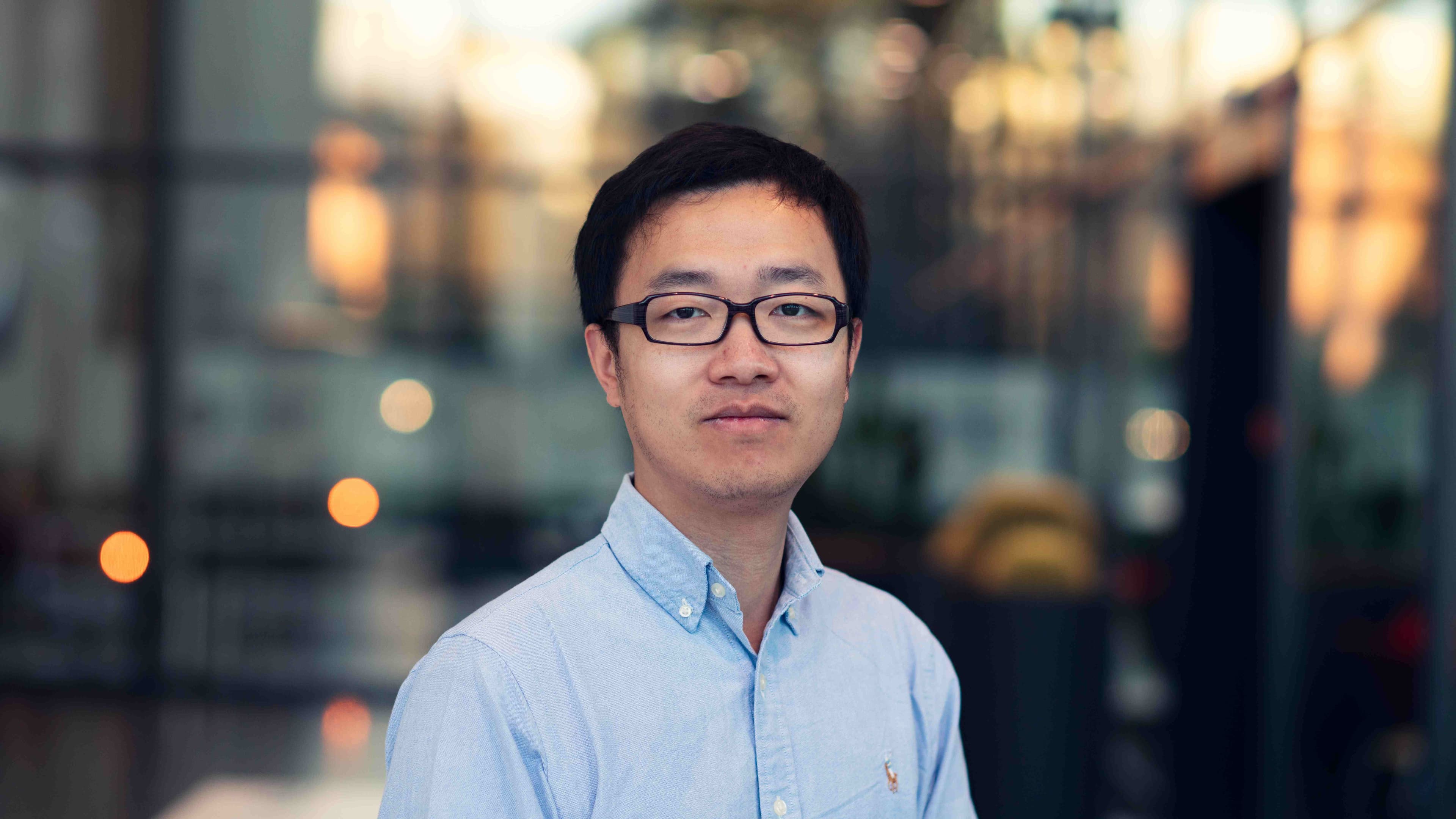 Lijun%20Liu001 Portrait of researcher Lijun Liu wearing glasses and a light blue shirt, standing in front of a blurred lab-like background.
